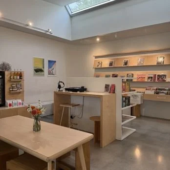 A cozy bookstore with wooden shelves filled with books, a small table with a vase of flowers, and artwork on the wall. There is a high ceiling with skylights.