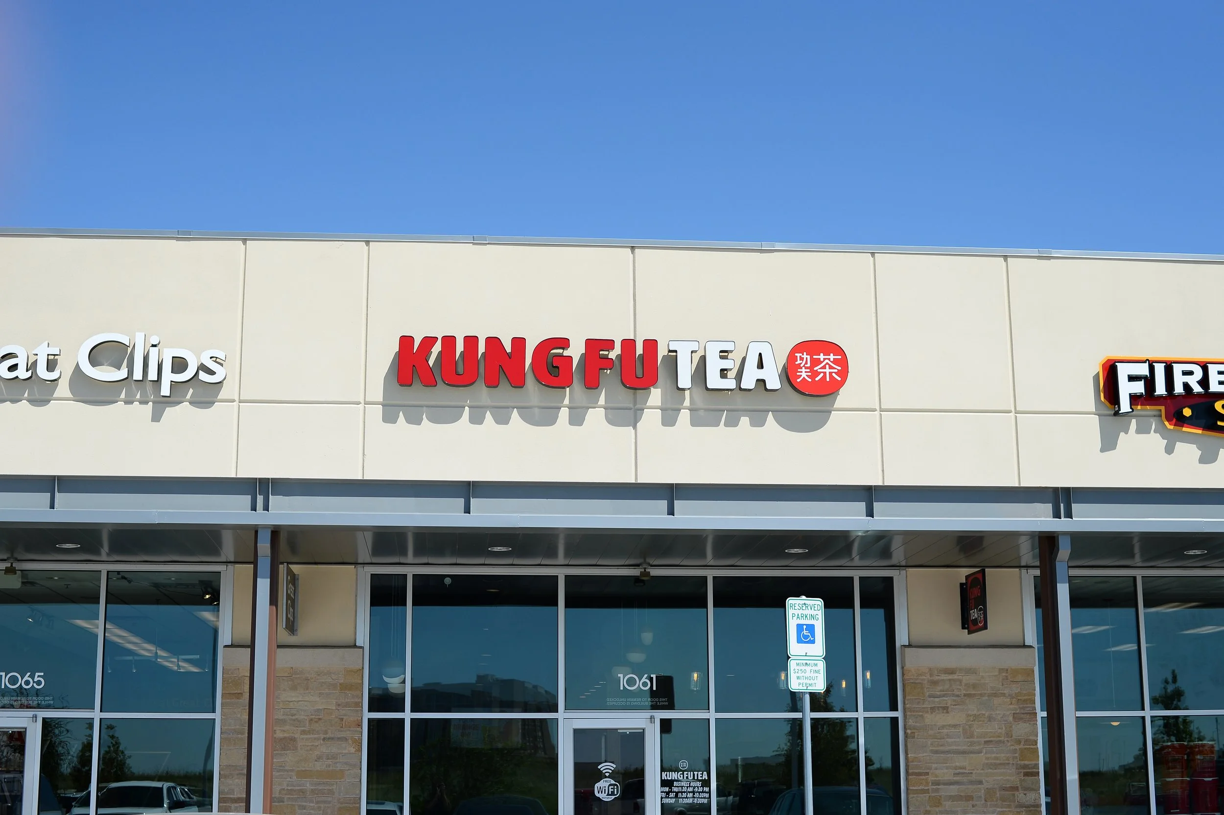 Storefront with signs for Great Clips, Kung Fu Tea, and Firehouse Subs, with a reserved parking sign and glass entrance doors, under a clear blue sky.