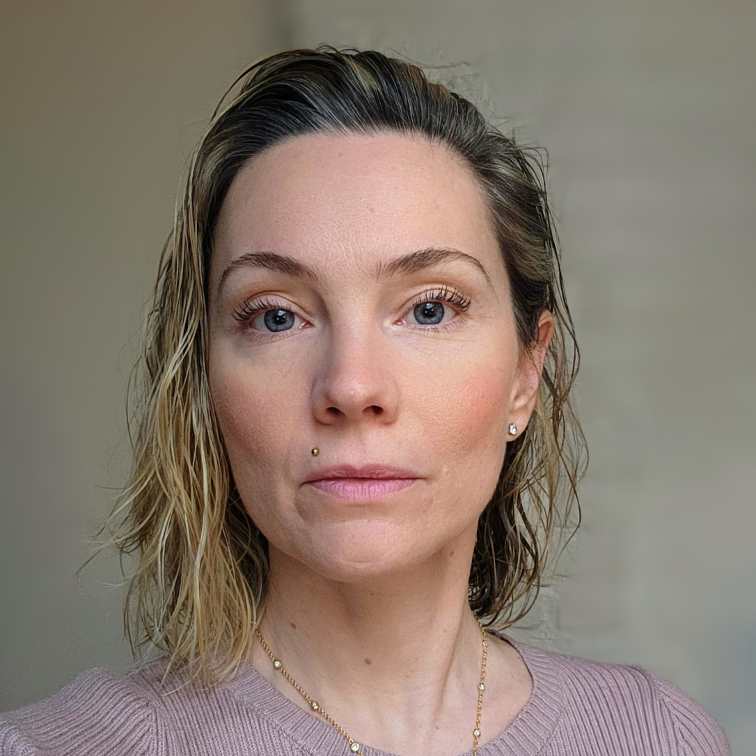 A blonde blue eyed woman wearing a lilac sweater in front of a blur background