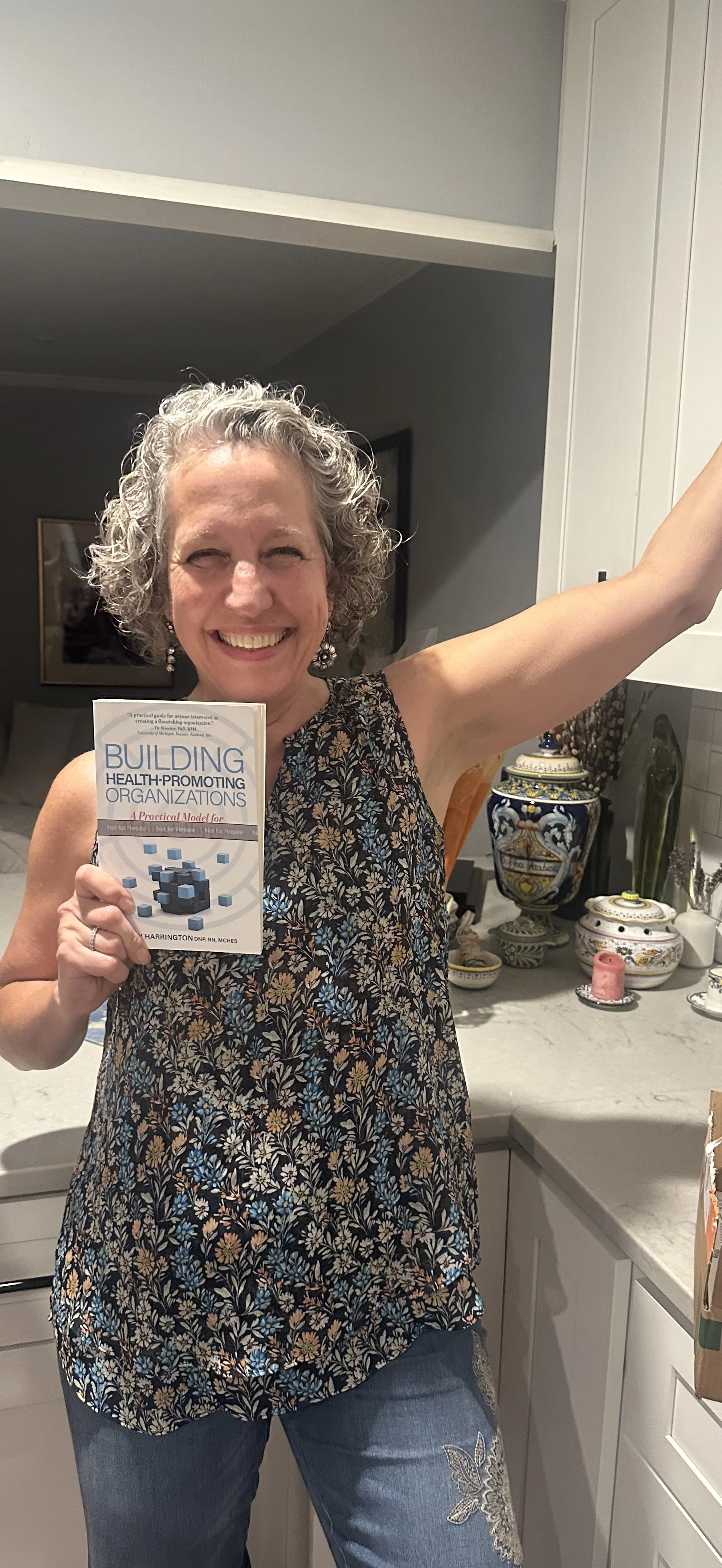 A woman with curly gray hair smiling and holding a book titled 'Building Health-Promoting Organizations'. She is standing in a kitchen with decorative plates and vases in the background, wearing a sleeveless, floral-patterned top and jeans.