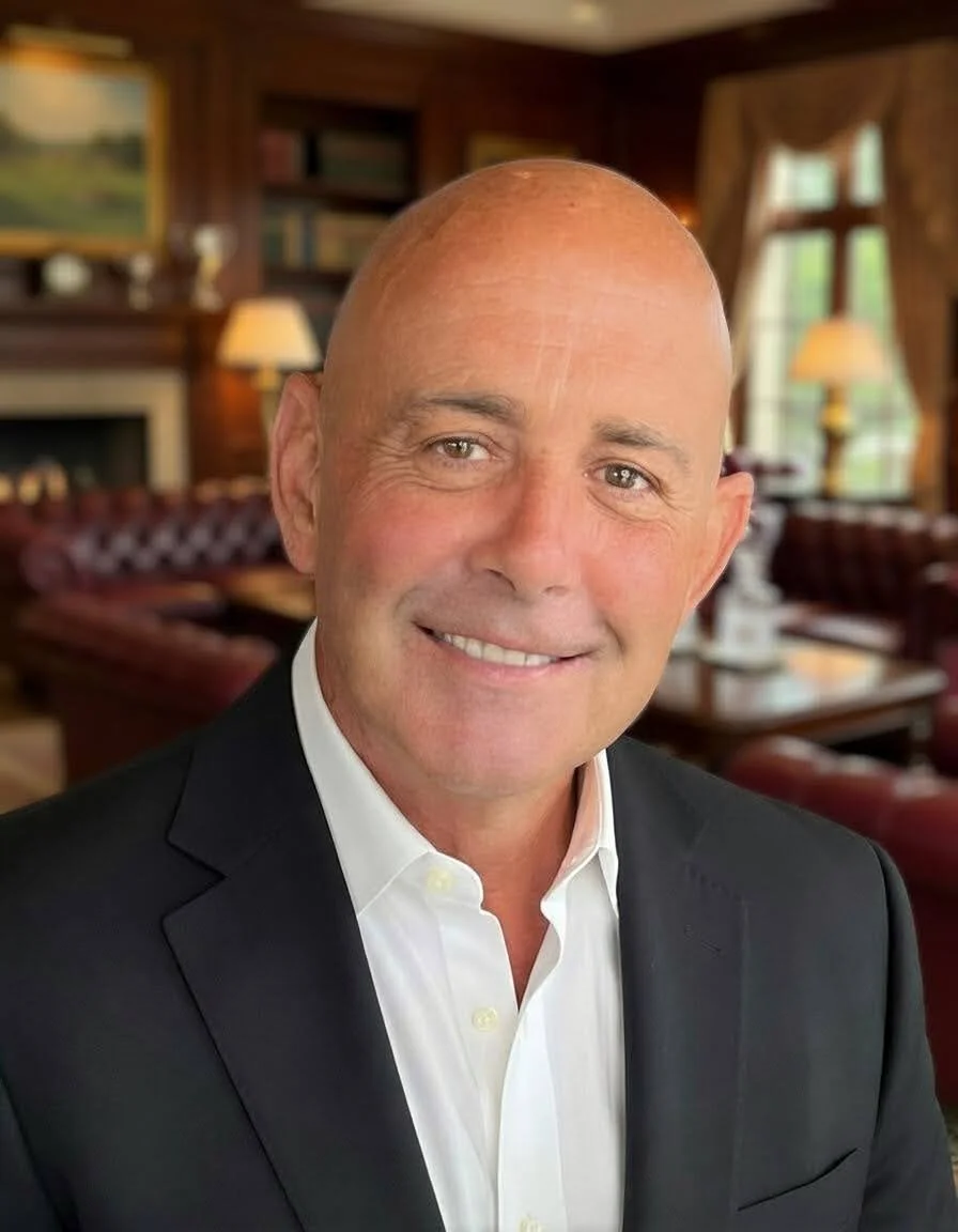 A smiling middle-aged man with a bald head, dressed in a white shirt and black blazer, in a cozy room with wooden walls, bookshelves, lamps, and a fireplace.
