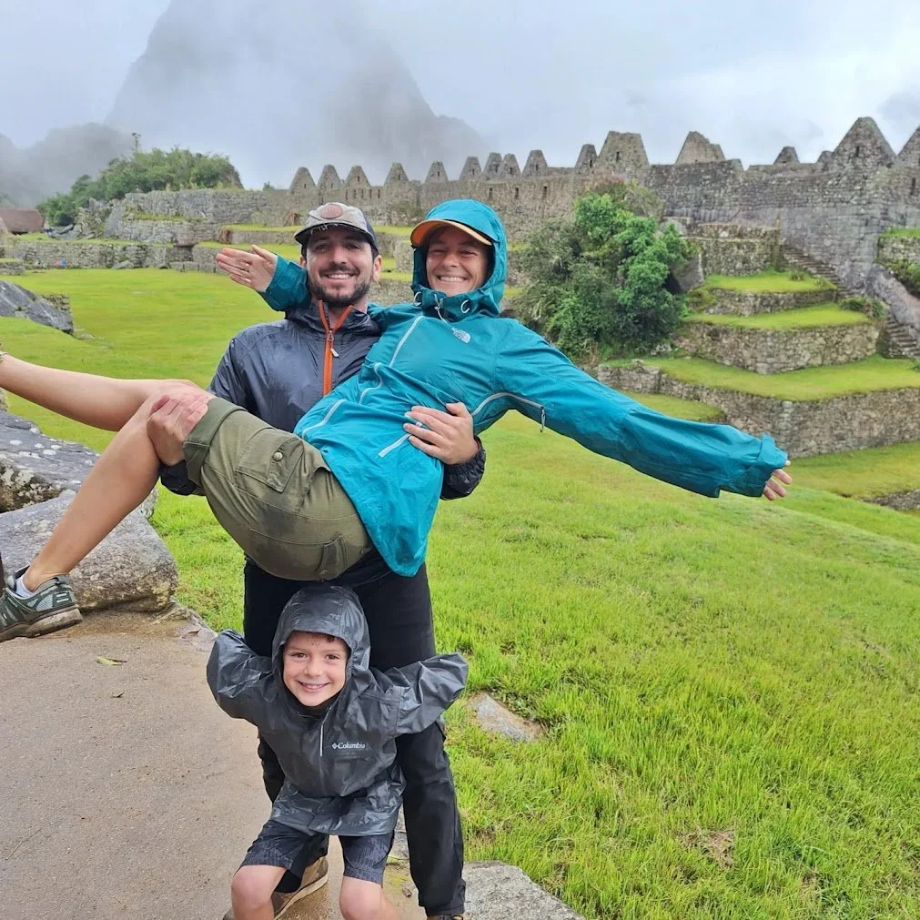 A family of three in rain jackets smiling at Machu Picchu, with ancient stone ruins and terraces in the background on a rainy day.