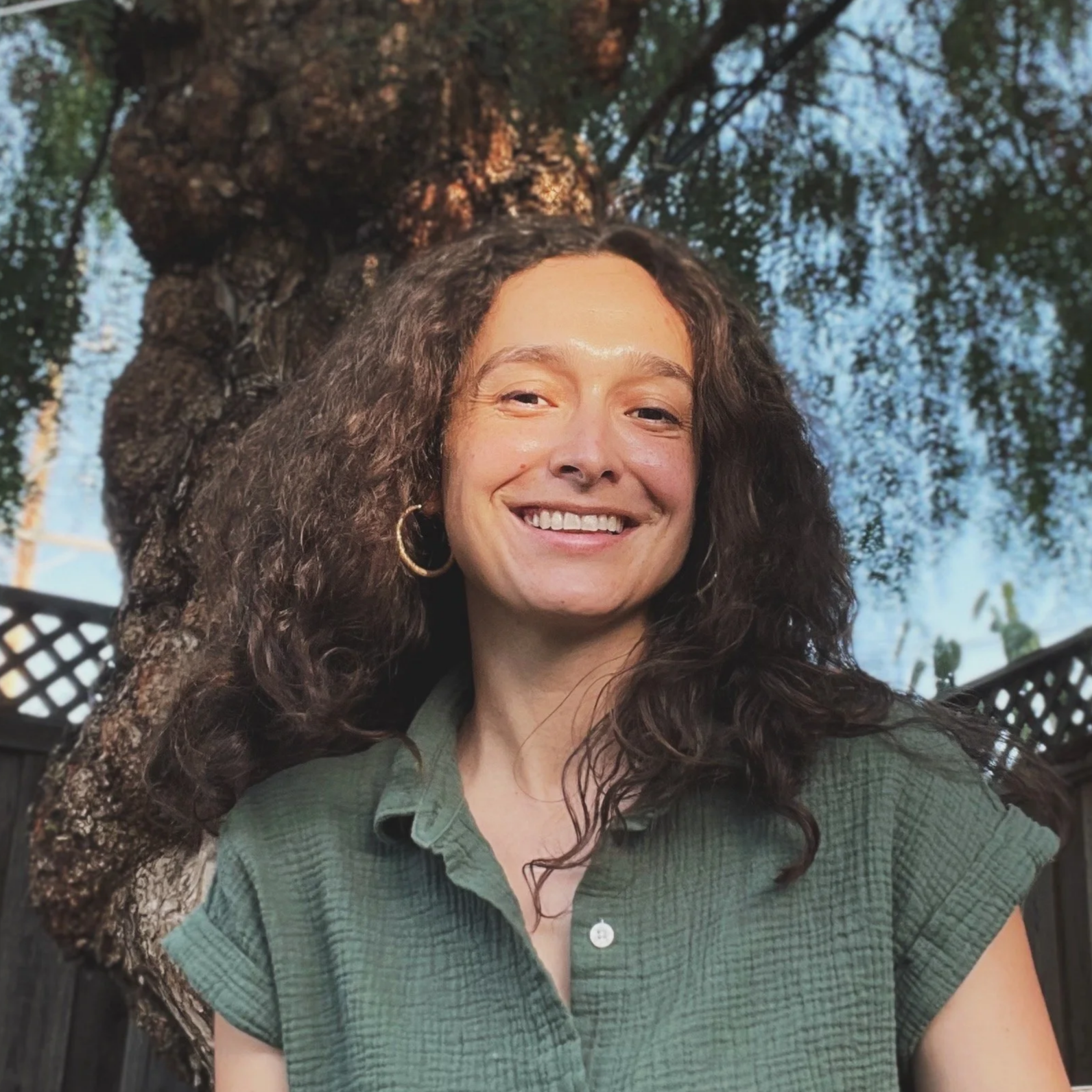Smiling woman with curly hair wearing a green shirt and hoop earrings outdoors near a tree with a fence in the background.