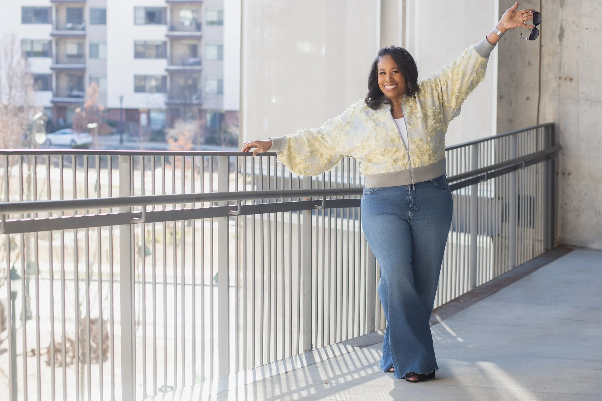 Smiling woman in a yellow and gray jacket and jeans standing on a balcony, holding sunglasses, with urban buildings in the background.