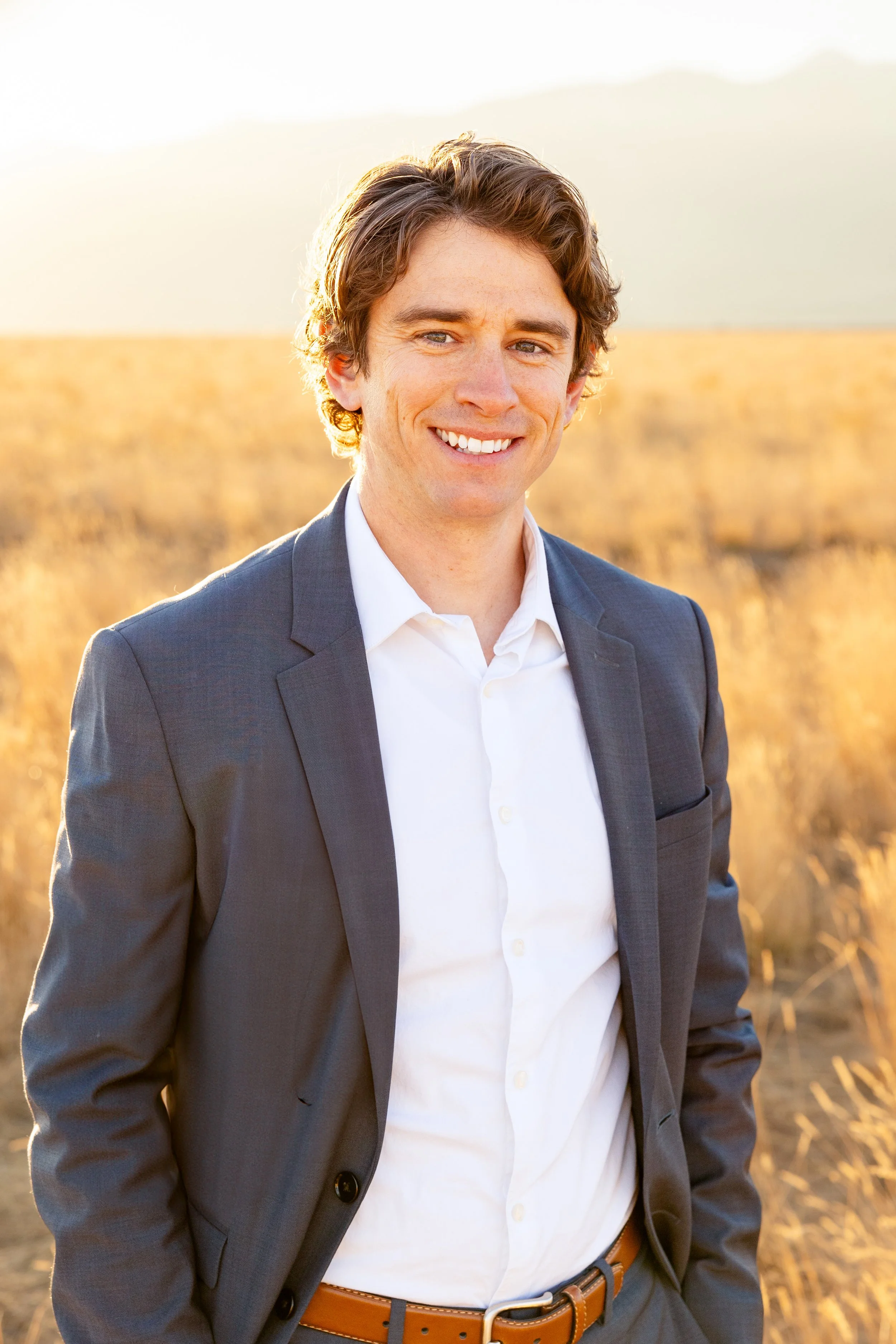 A man with brown hair and blue eyes in a gray suit and white shirt standing outdoors in a field at sunset, smiling at the camera.