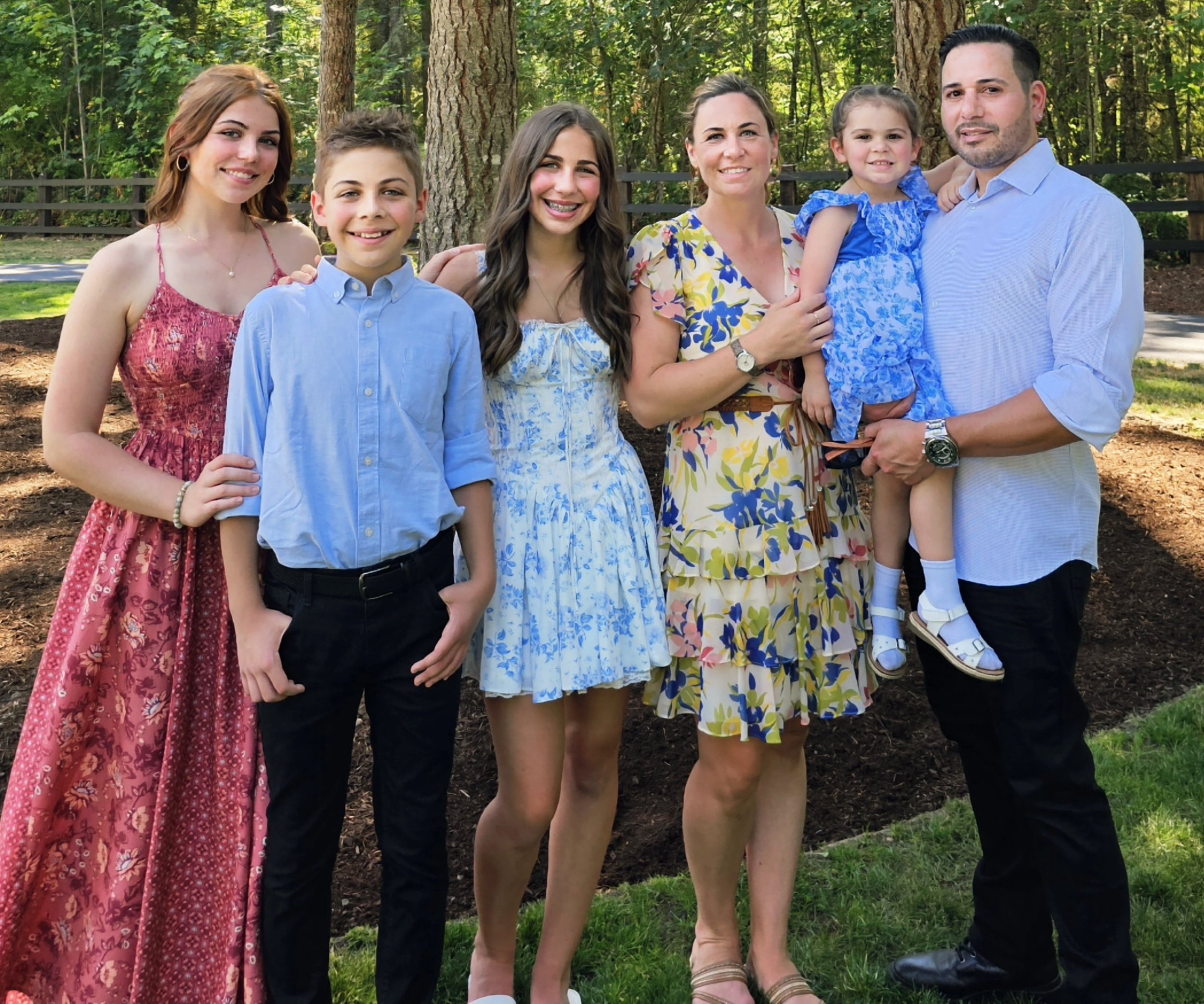 Family of six standing outdoors in a park with trees and a wooden fence, smiling for a group photo.