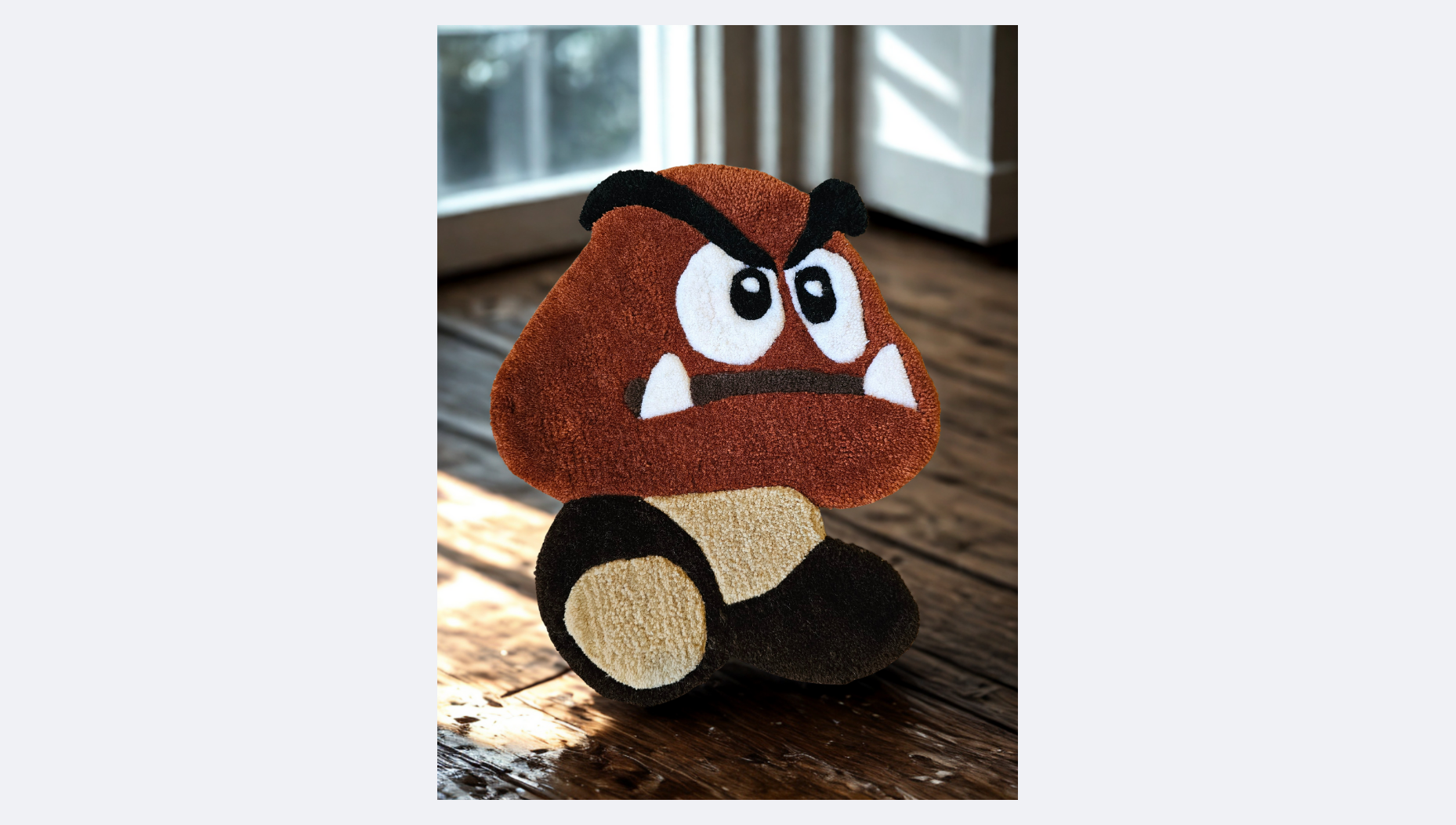 A plush toy resembling a brown, angry-looking Goomba from Super Mario, with big eyes, eyebrows, white teeth, and black shoes, sitting on a wooden floor near a window.