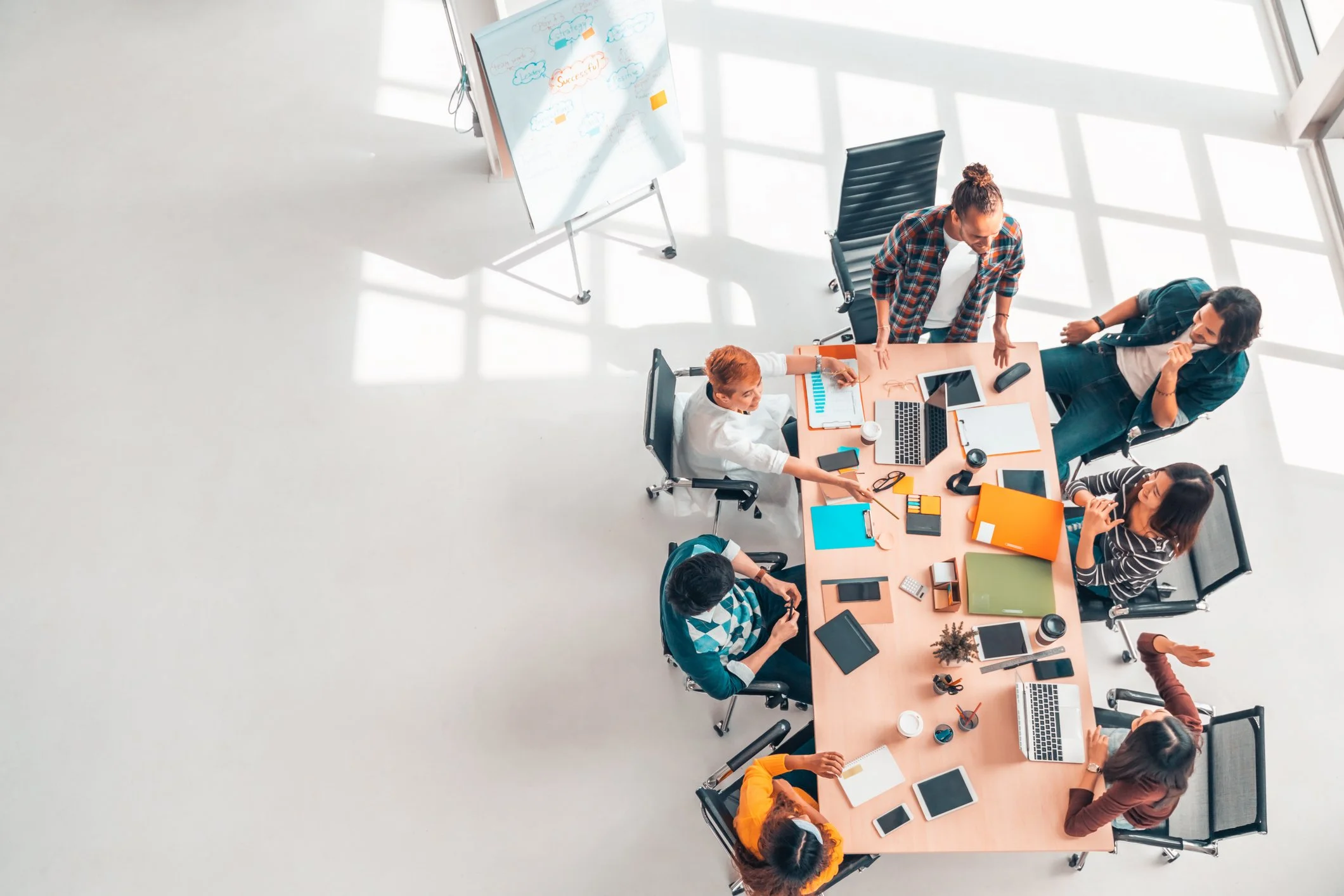 Overhead view of a diverse group of seven people sitting and standing around a large conference table in a bright office, engaged in a meeting with laptops, tablets, notebooks, and office supplies on the table.