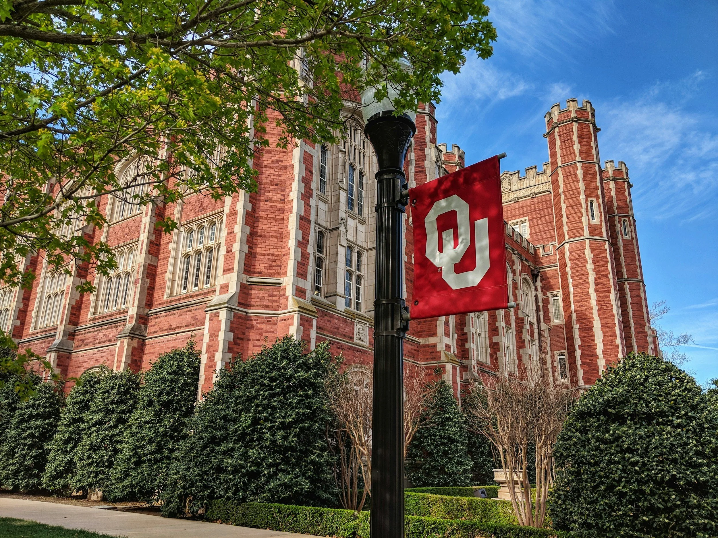 Red brick university building with tall windows, surrounded by trees and bushes, with a red flag displaying the University of Oklahoma logo on a black lamp post in the foreground and a blue sky with wispy clouds in the background.