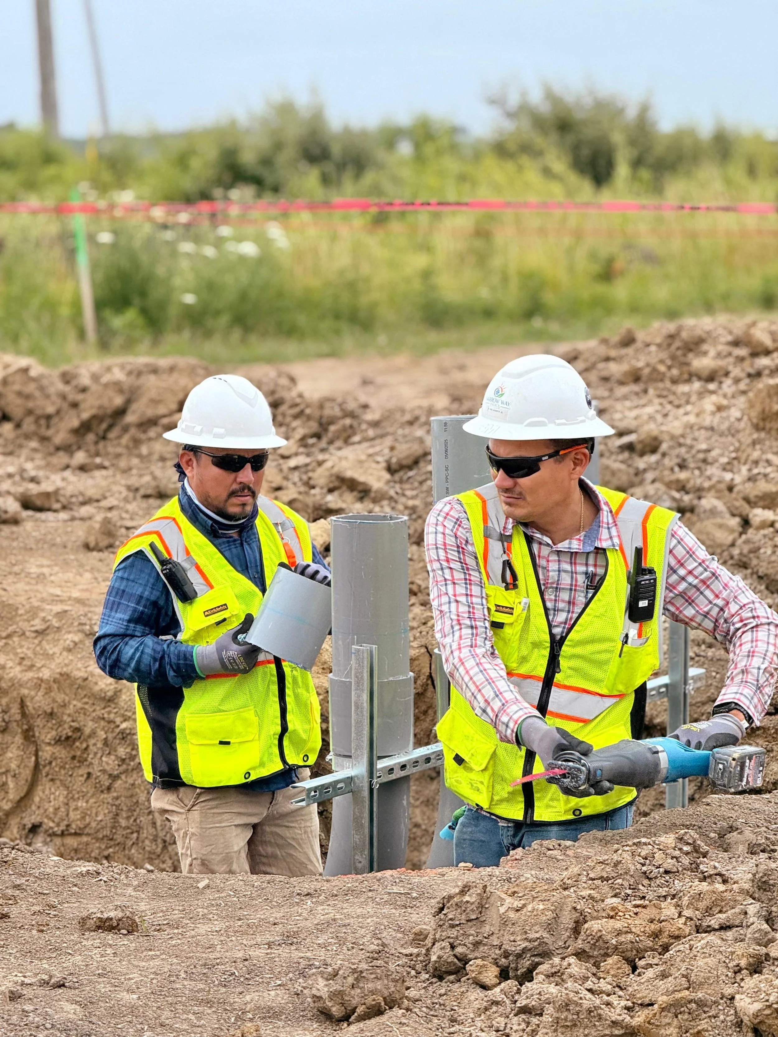 Two construction workers wearing safety vests, helmets, and sunglasses working on a utility installation at a construction site with dirt and pipes.