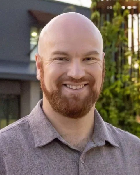 Headshot of a smiling man with a beard and shaved head, wearing a gray collared shirt, outdoors in front of a building and greenery.