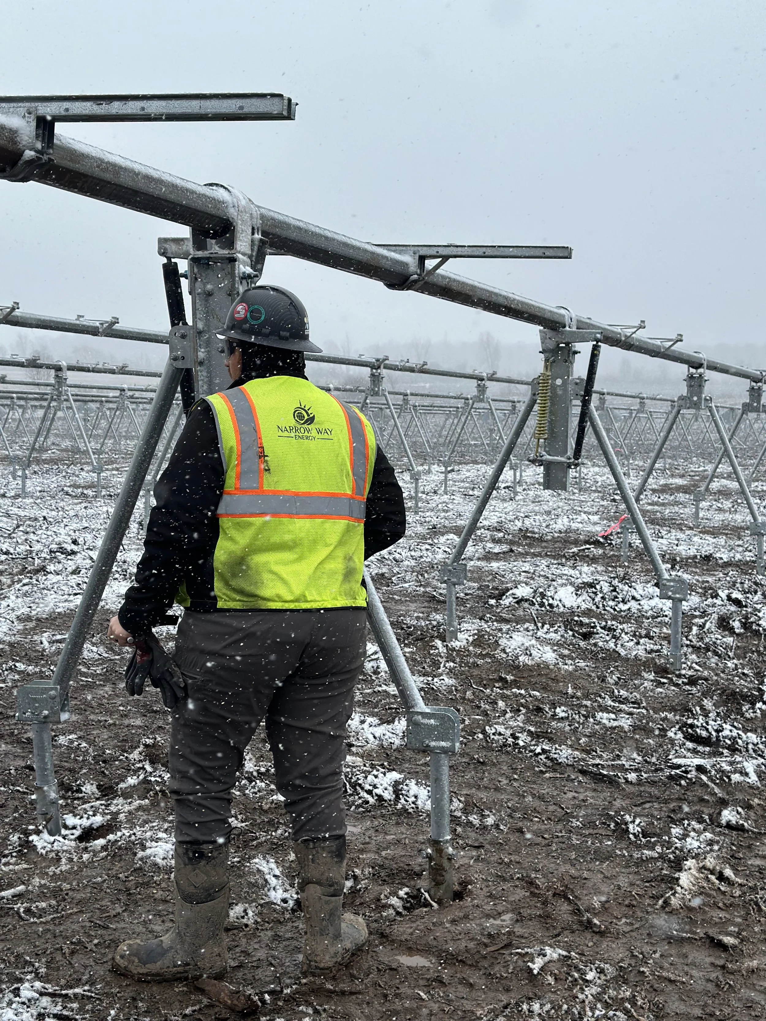 A worker in a high-visibility safety vest and helmet inspecting solar panel mounting structures in a snowy field.