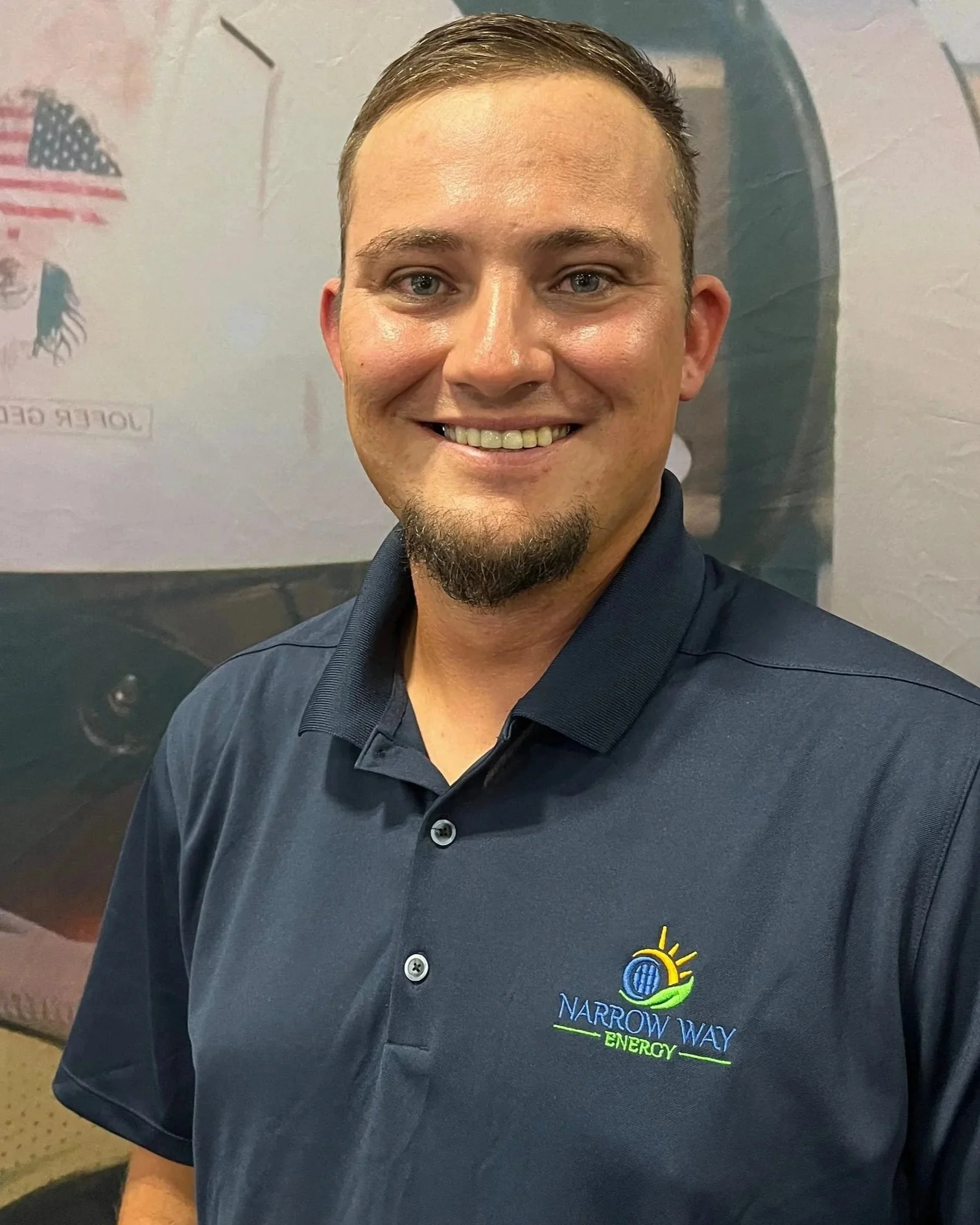 A smiling man in a navy blue polo shirt with "Narrow Way Energy" logo stands in front of a wall with American flag and energy-related images.