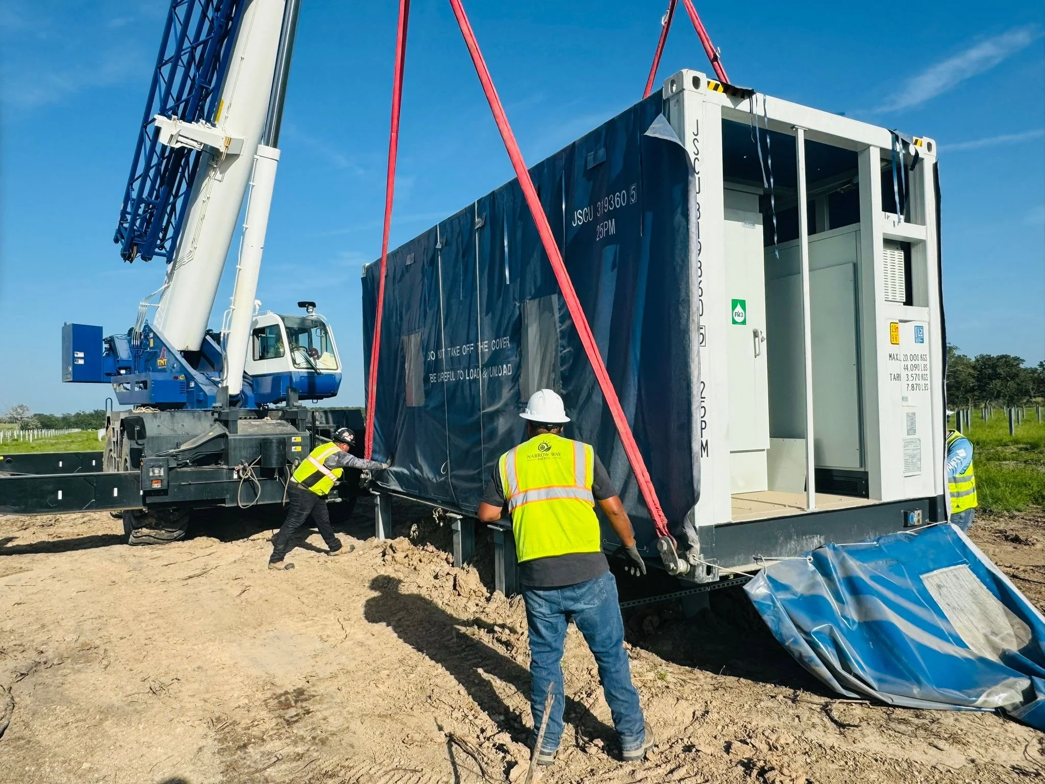 Workers in safety vests and helmets are operating a crane to place a modular unit onto a construction site with a clear blue sky background.