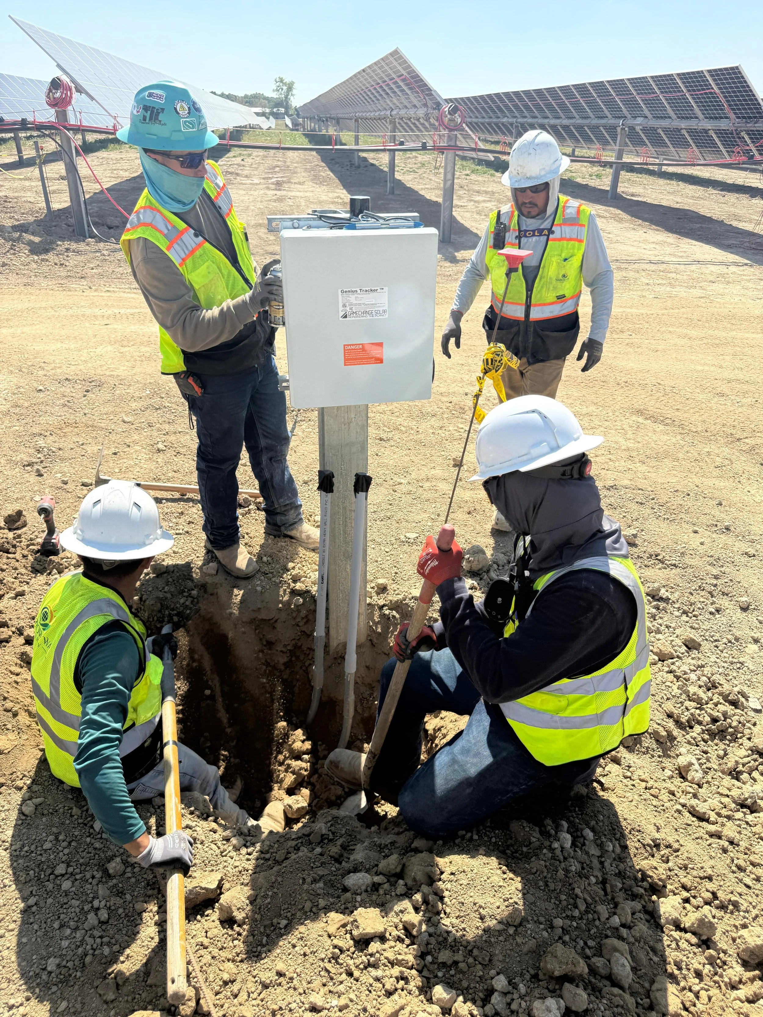 Four construction workers wearing helmets and reflective vests working on an electrical or solar power system installation outdoors, with solar panels in the background.