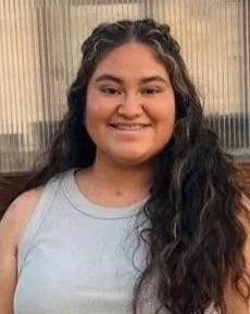 Young woman with long curly hair smiling, wearing a light gray sleeveless top, standing in front of a window with vertical blinds.
