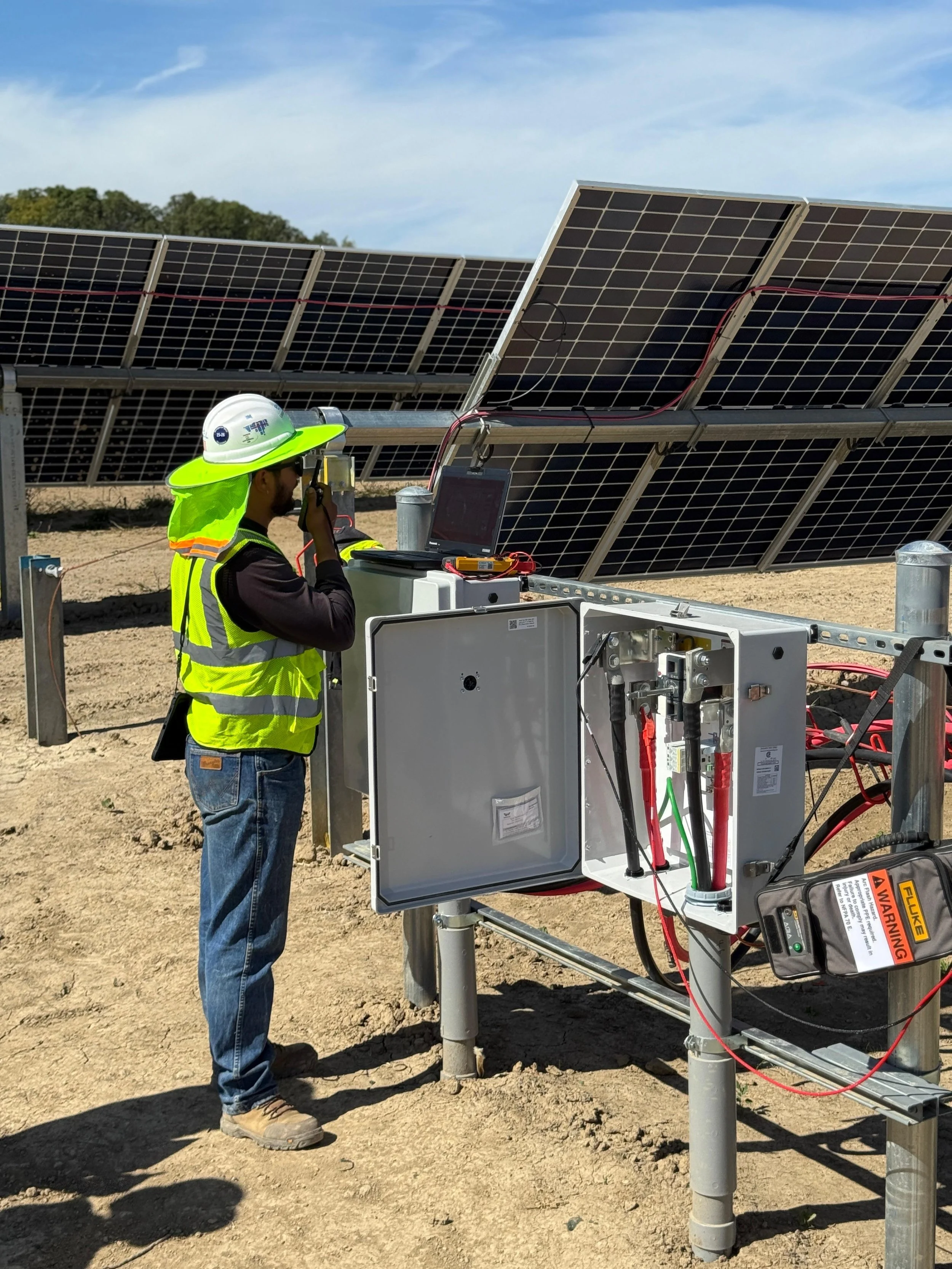 A worker in a yellow safety vest, white hard hat, and jeans is inspecting electric equipment at a solar panel installation site on a sunny day.