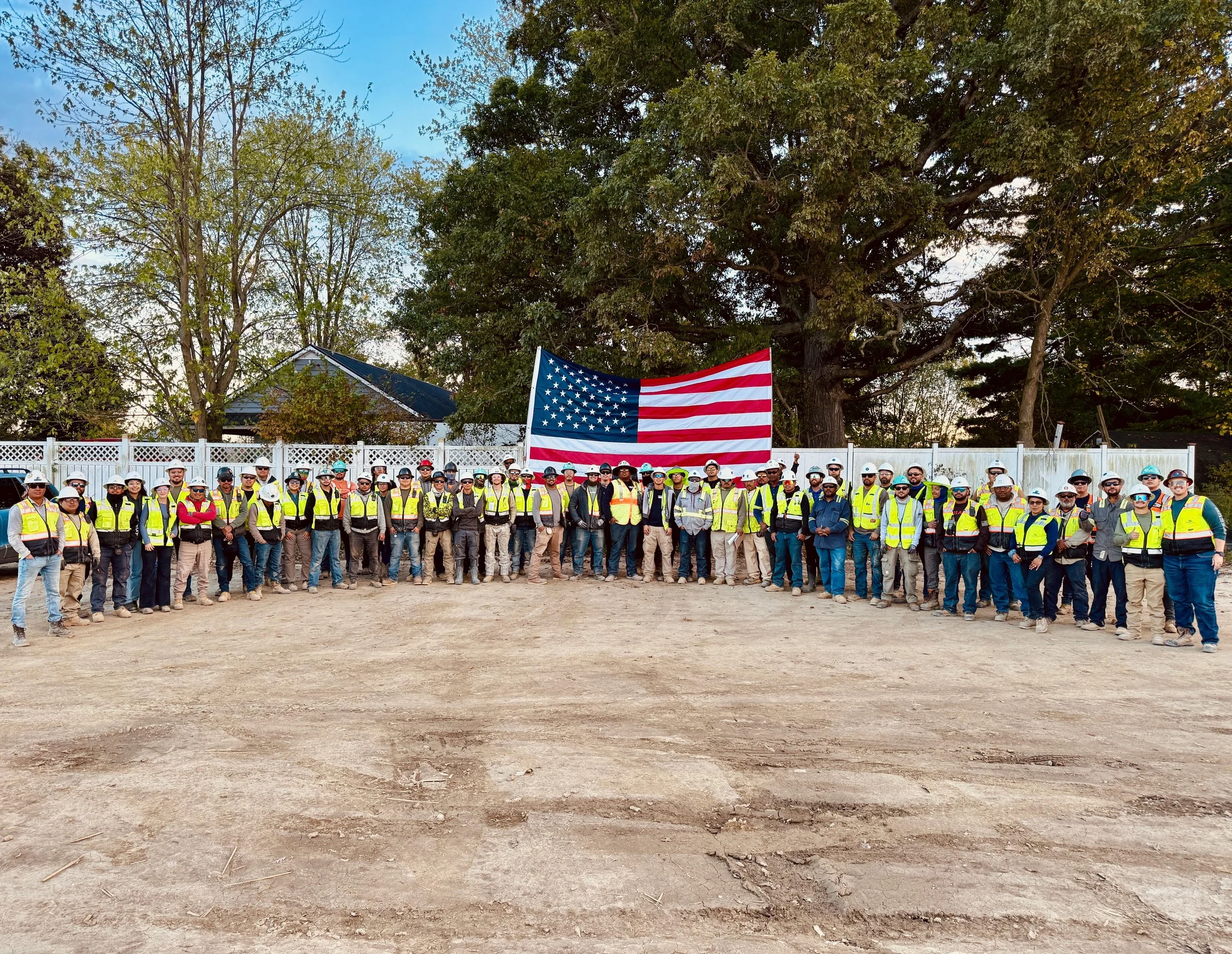 Group of construction workers standing outdoors in front of a white fence, wearing safety vests and helmets, with an American flag hung on the fence behind them, trees and houses in the background.