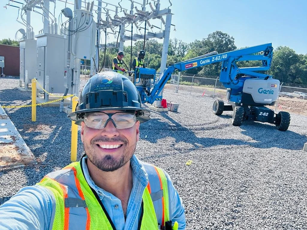 A construction worker in safety gear smiling at the camera on a gravel site with electrical equipment and a person in a lift in the background.