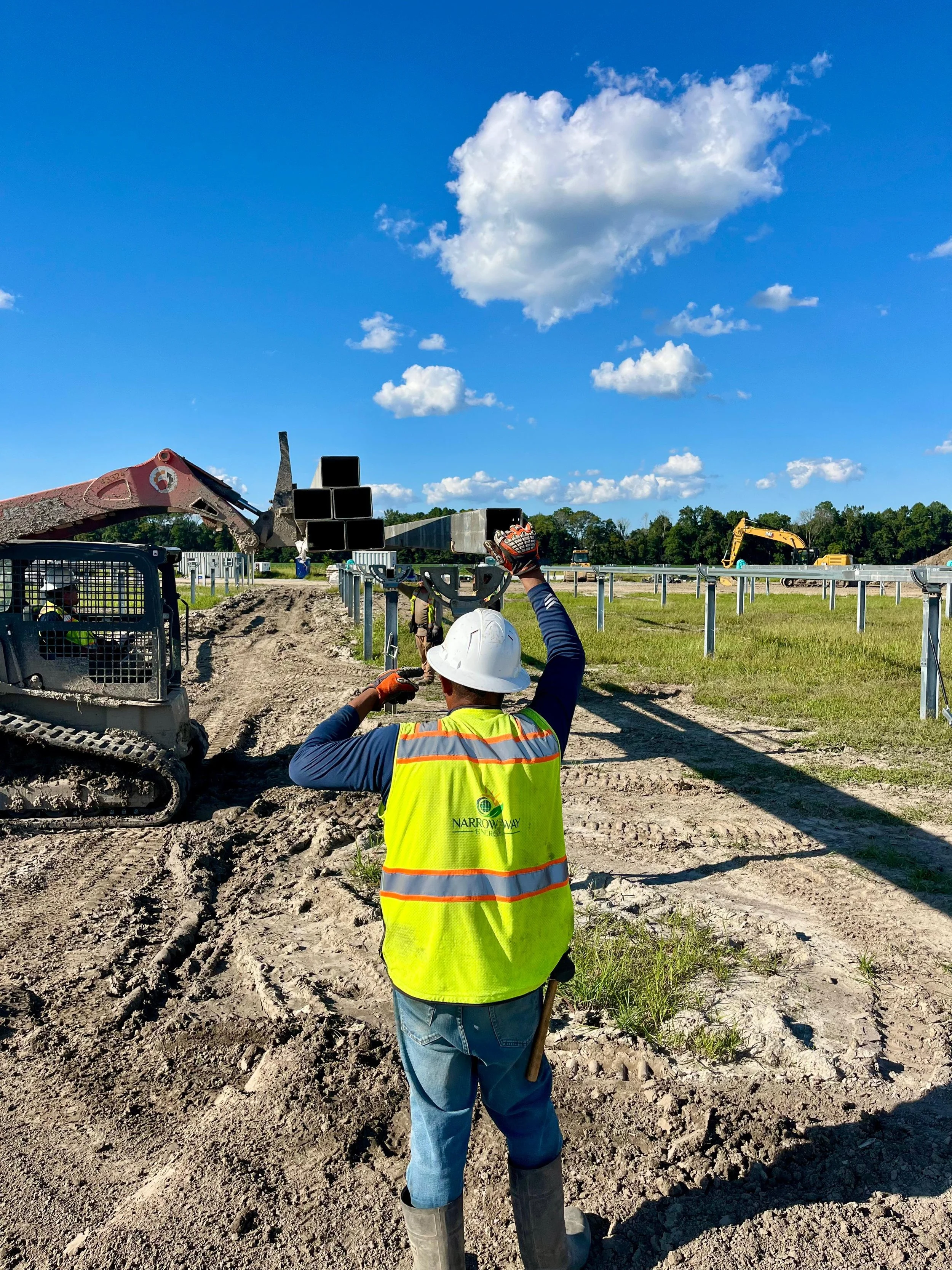 Construction worker in a safety vest and hard hat working on a construction site with excavators and steel construction supports under a blue sky with clouds.