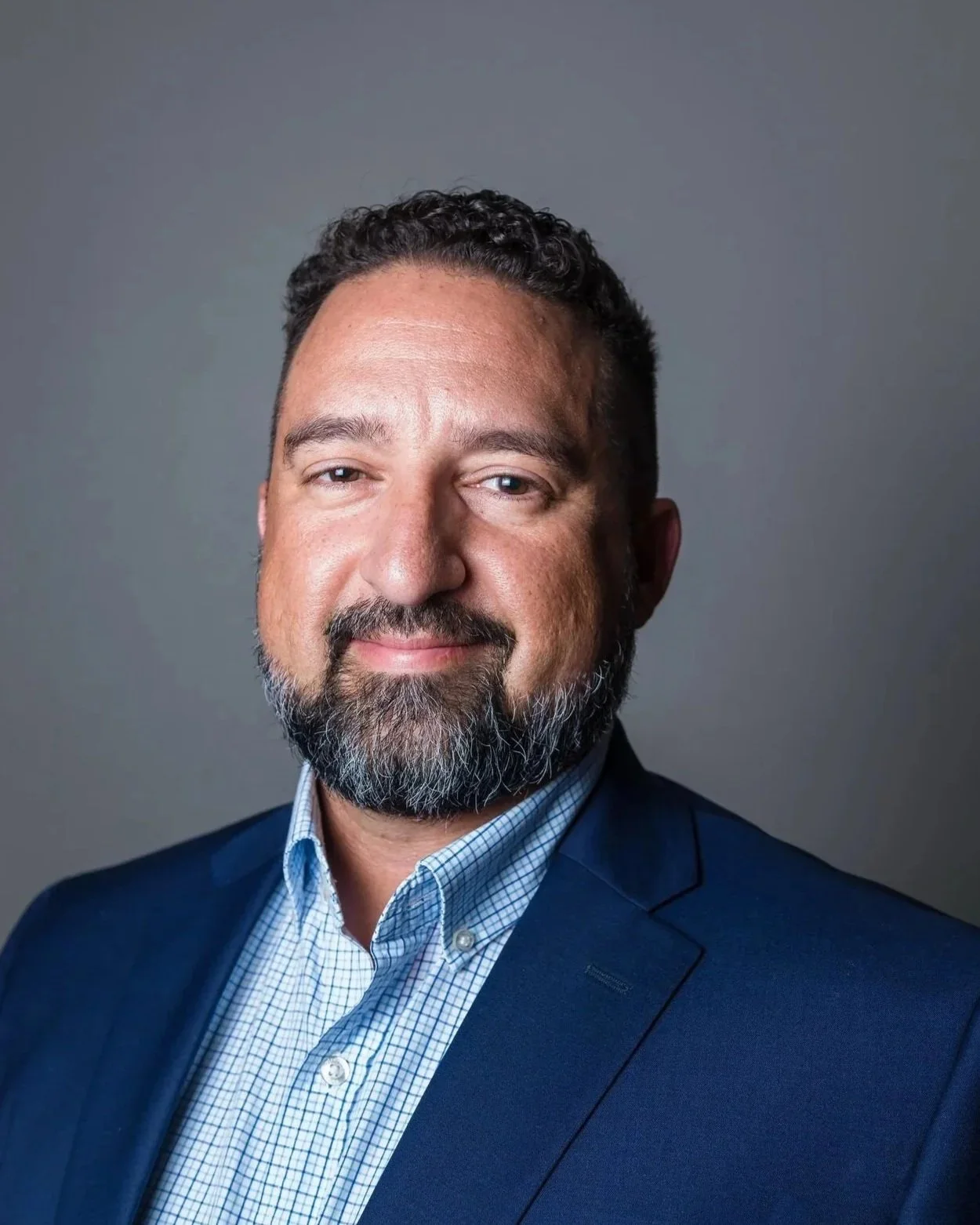 Portrait of a middle-aged man with dark curly hair and a gray beard, wearing a blue blazer and a checkered dress shirt, smiling slightly, against a gray background.