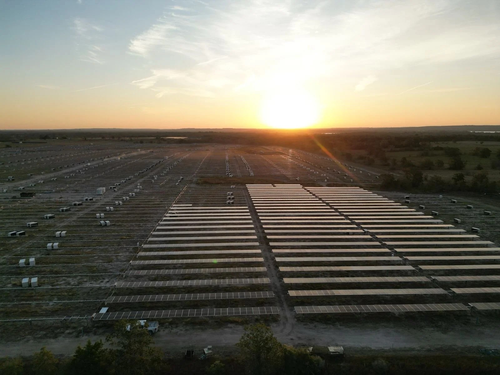 A large solar panel farm with numerous solar panels and some parked vehicles at sunrise or sunset.