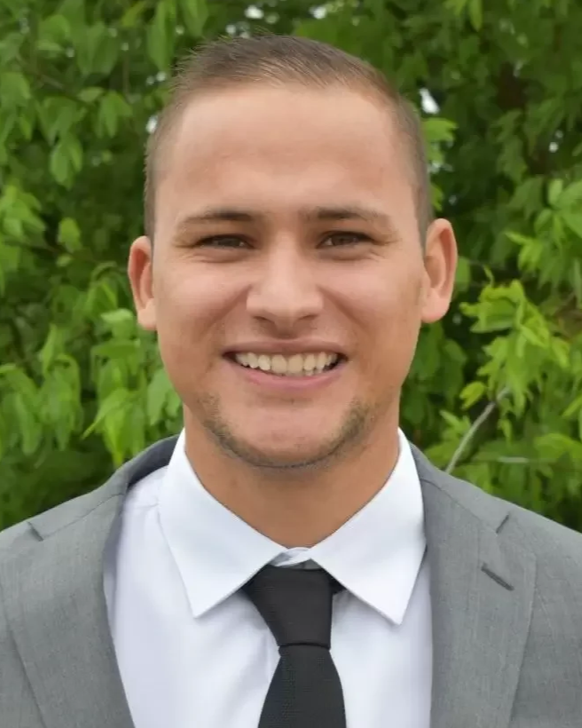 Close-up of a smiling young man in a gray suit, white shirt, and black tie, standing outdoors with green leafy trees in the background.