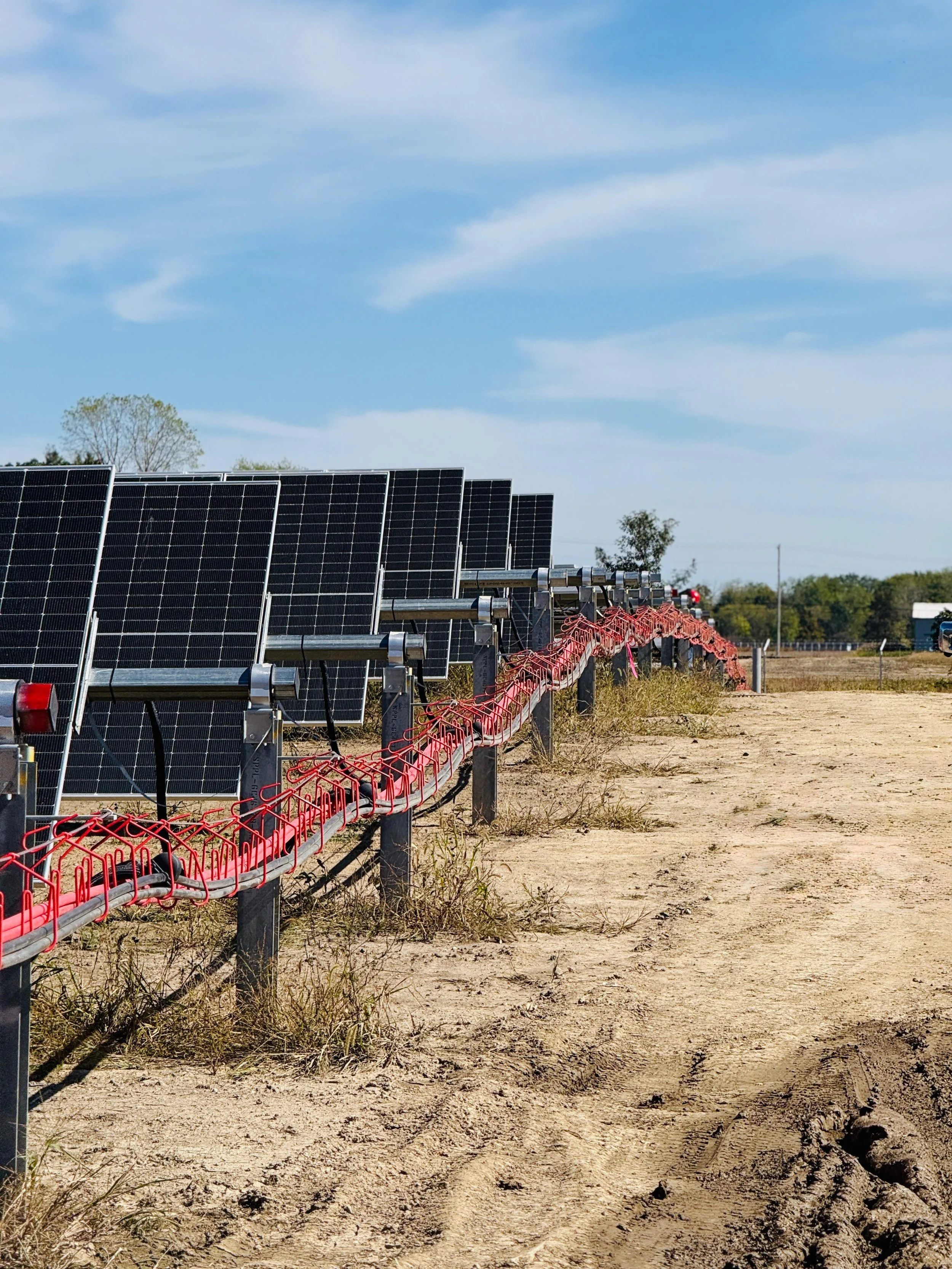 Solar panels mounted on metal frames in a field under a blue sky with scattered clouds, with tire tracks and some grass on the dirt ground.