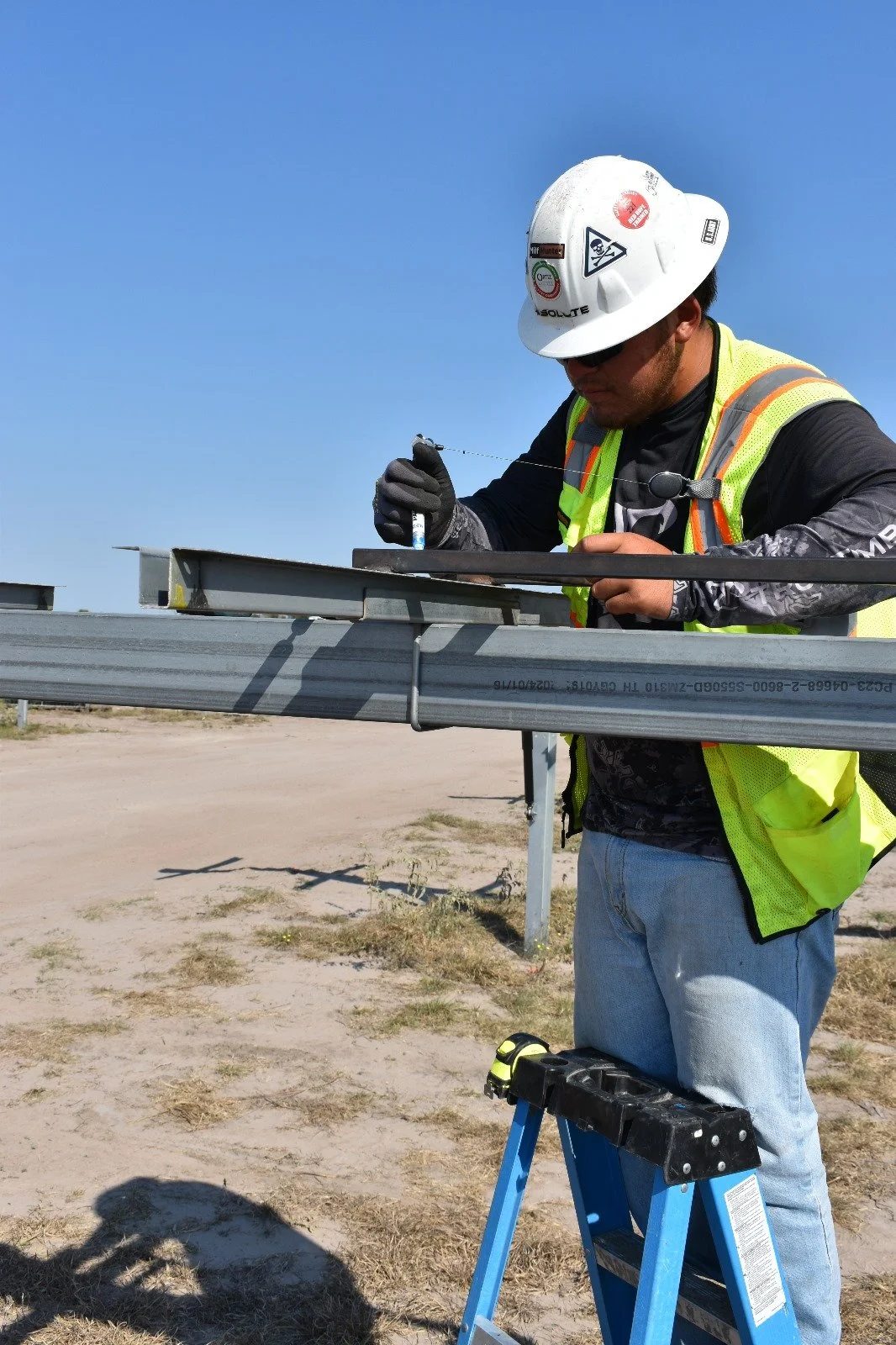 A construction worker wearing a safety helmet and vest is working outdoors on a metal structure, measuring or marking it with a marker. The sky is clear and sunny.