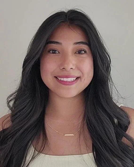 A young woman with long dark hair, smiling, wearing a beige top and a gold necklace.