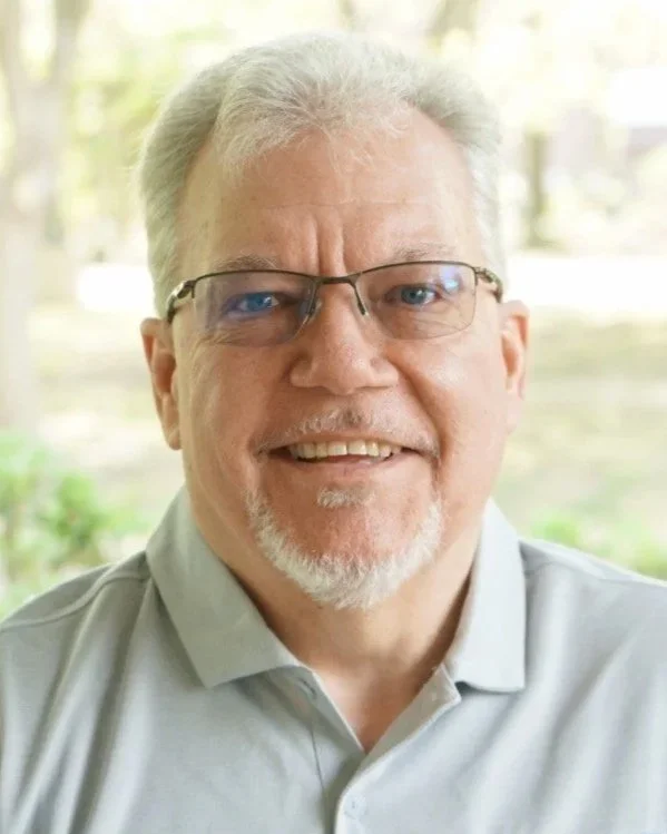 Close-up of a smiling middle-aged man with gray hair and beard, wearing glasses and a gray collared shirt, in a bright indoor setting.