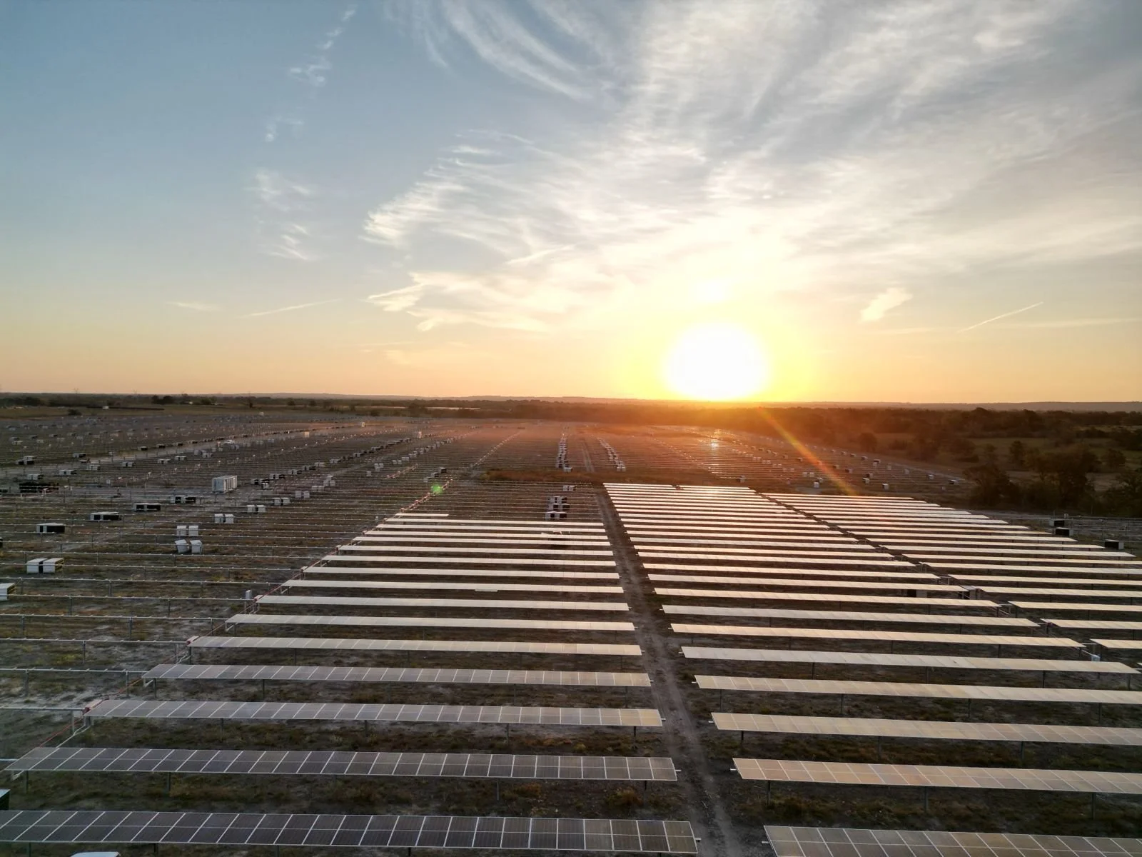 A large solar farm with numerous solar panels arranged in rows, captured at sunset with the sun low on the horizon and a partly cloudy sky.
