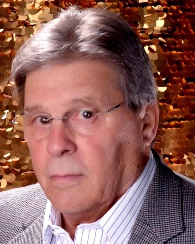 Close-up of a mature man with gray hair, wearing glasses, a white striped shirt, and a checkered blazer, standing in front of a gold sequin backdrop.