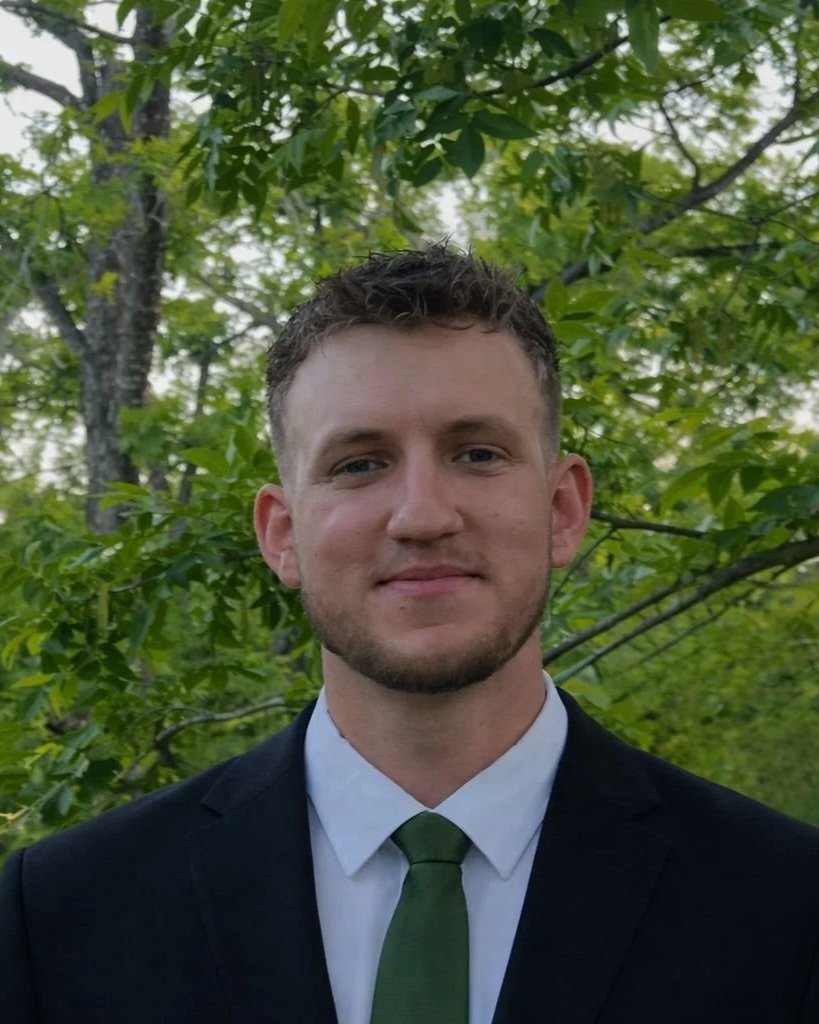 A young man with short curly hair and a beard, wearing a dark suit, white shirt, and green tie, standing outdoors with green trees in the background.