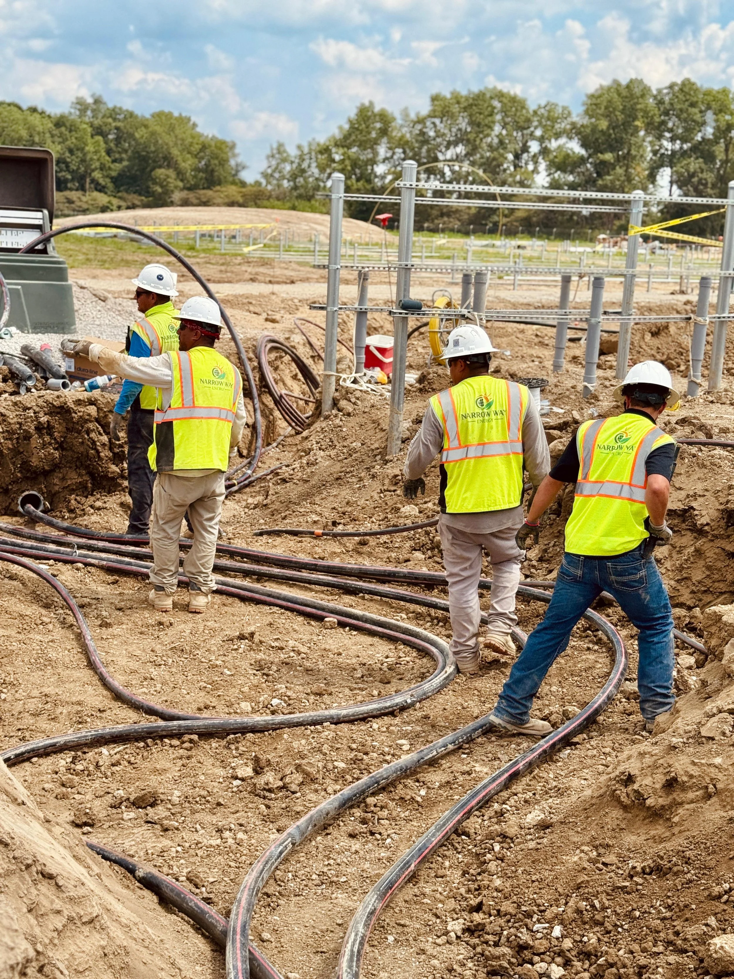 Construction workers wearing safety vests and helmets working on underground wiring or piping on a construction site, with dirt and equipment around.