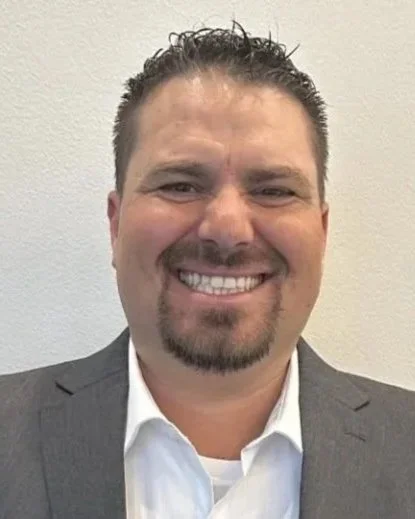 Smiling man with short dark hair, a trimmed beard and mustache, wearing a gray suit and white shirt, standing in front of a plain wall.