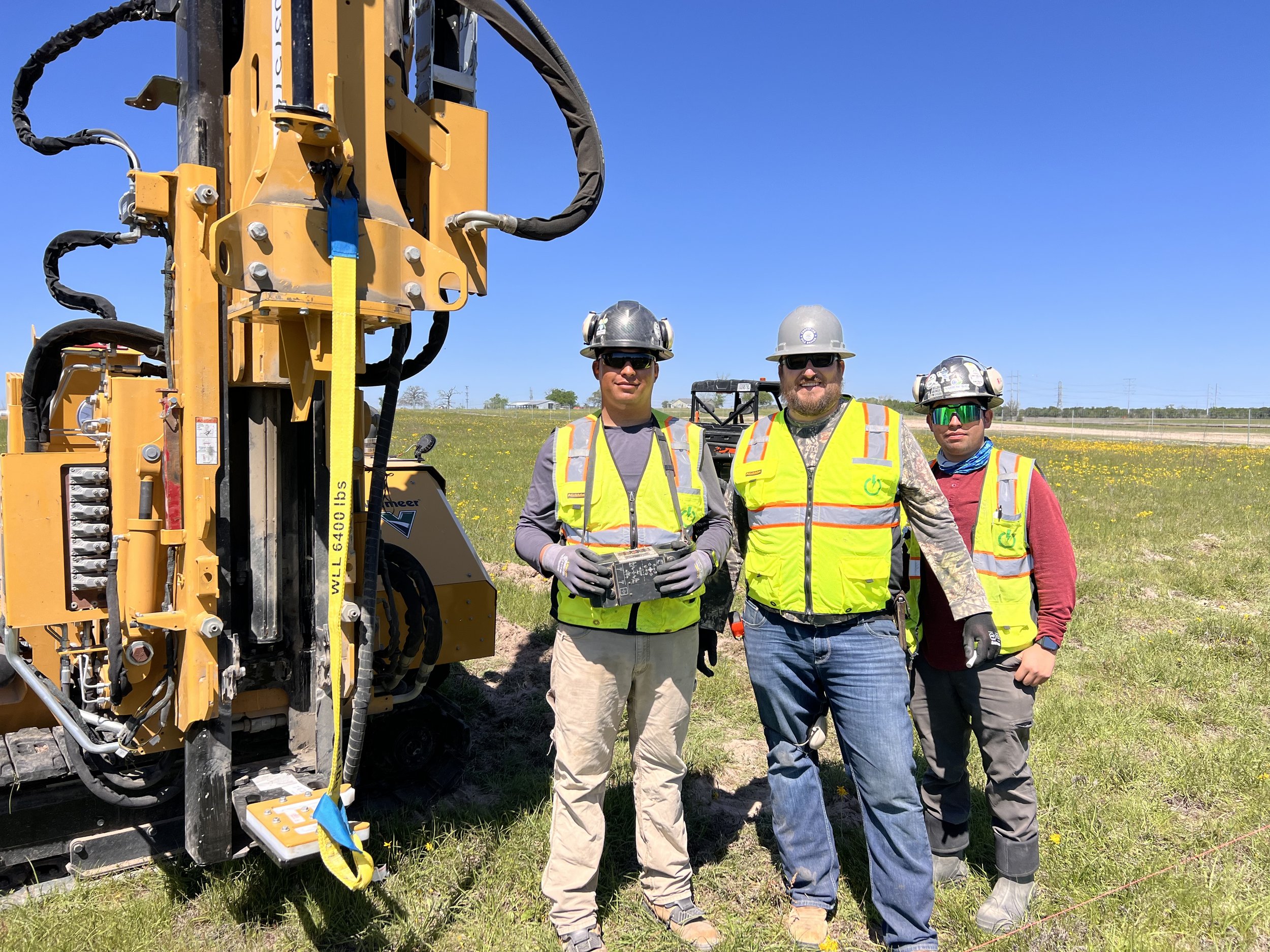Three construction workers wearing helmets and safety vests standing next to a large yellow drilling machine in an open field under a clear blue sky.