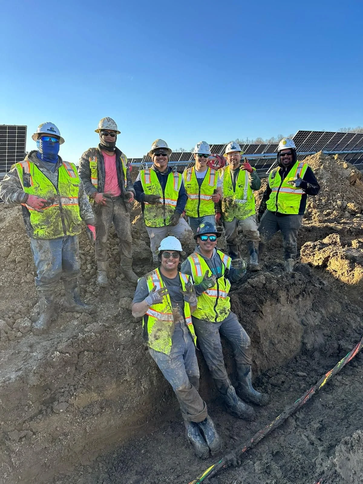 Group of construction workers wearing safety gear, hard hats, and reflective vests posing for a photo at a solar power site with solar panels visible in the background.