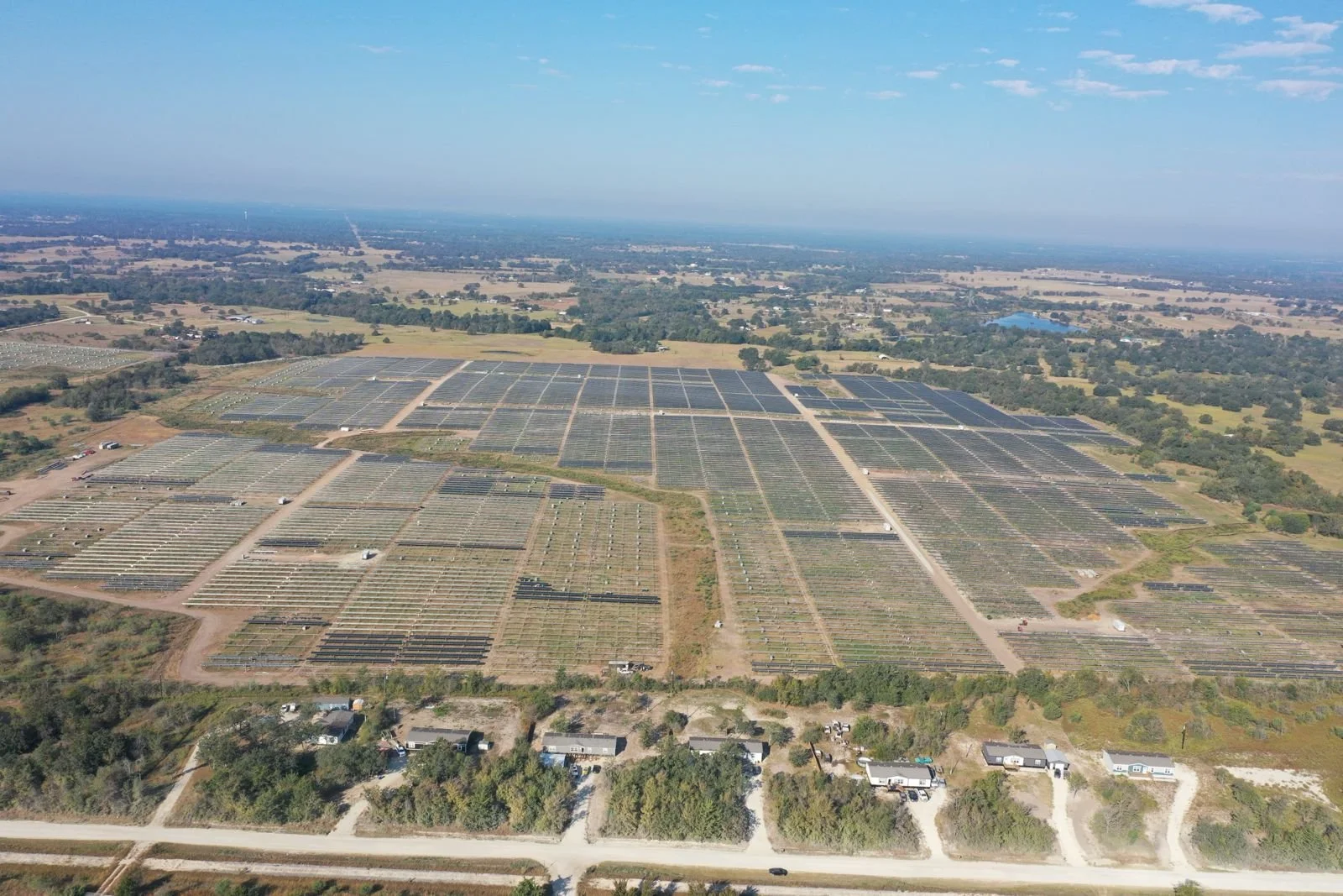 Aerial view of a large solar farm with numerous solar panels arranged in rows, surrounded by fields and sparse trees, under a partly cloudy sky.