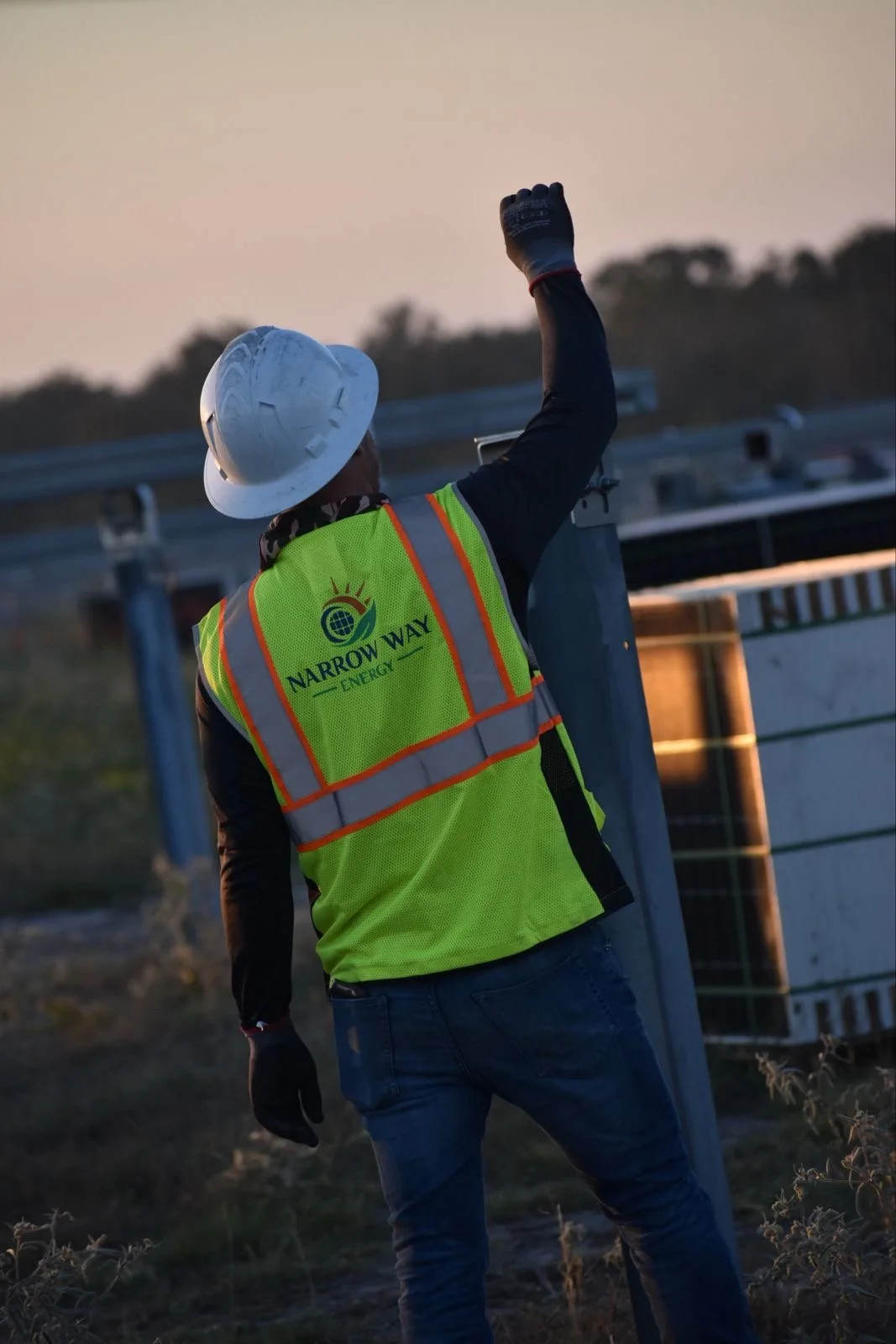 A construction worker wearing a yellow safety vest with the words "Narrow Way Energy," a white safety helmet, and gloves, standing outdoors at dusk, raising one fist in the air near industrial equipment.