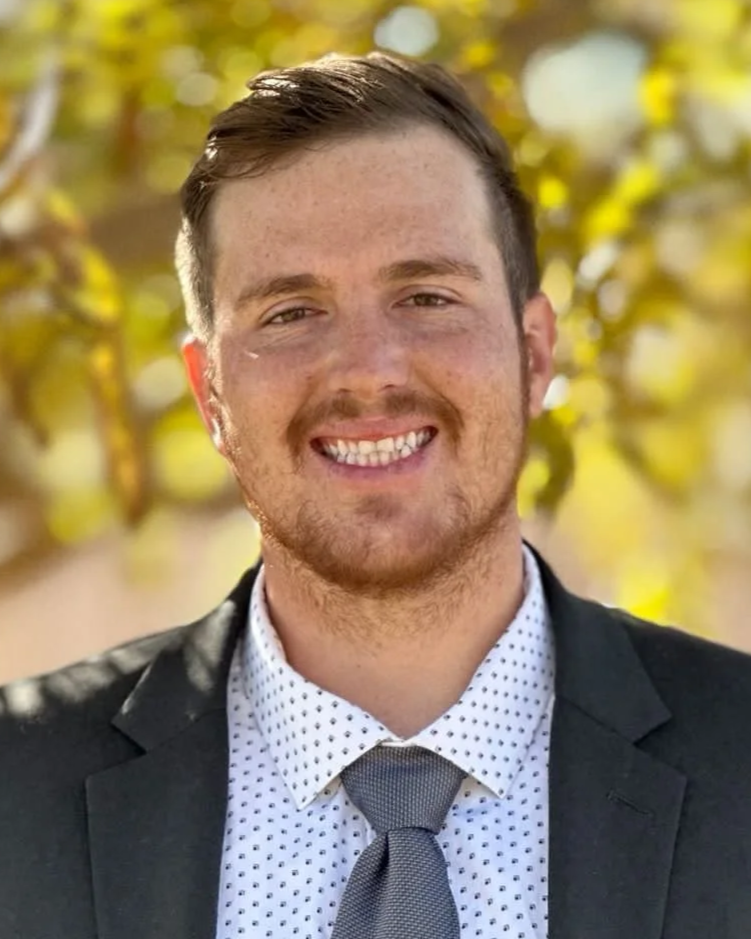 A young man with light brown hair, a beard, and a smile, wearing a dark suit, white shirt with small black polka dots, and a gray tie, standing outdoors with blurred green and yellow leaves in the background.
