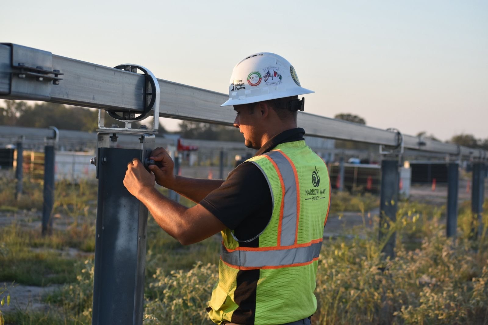 A worker in a yellow safety vest and a white hard hat adjusting equipment at an outdoor energy site during daytime.