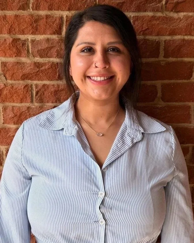 A woman with dark hair and a nose piercing smiling in front of a brick wall, wearing a light blue and white striped button-up shirt.