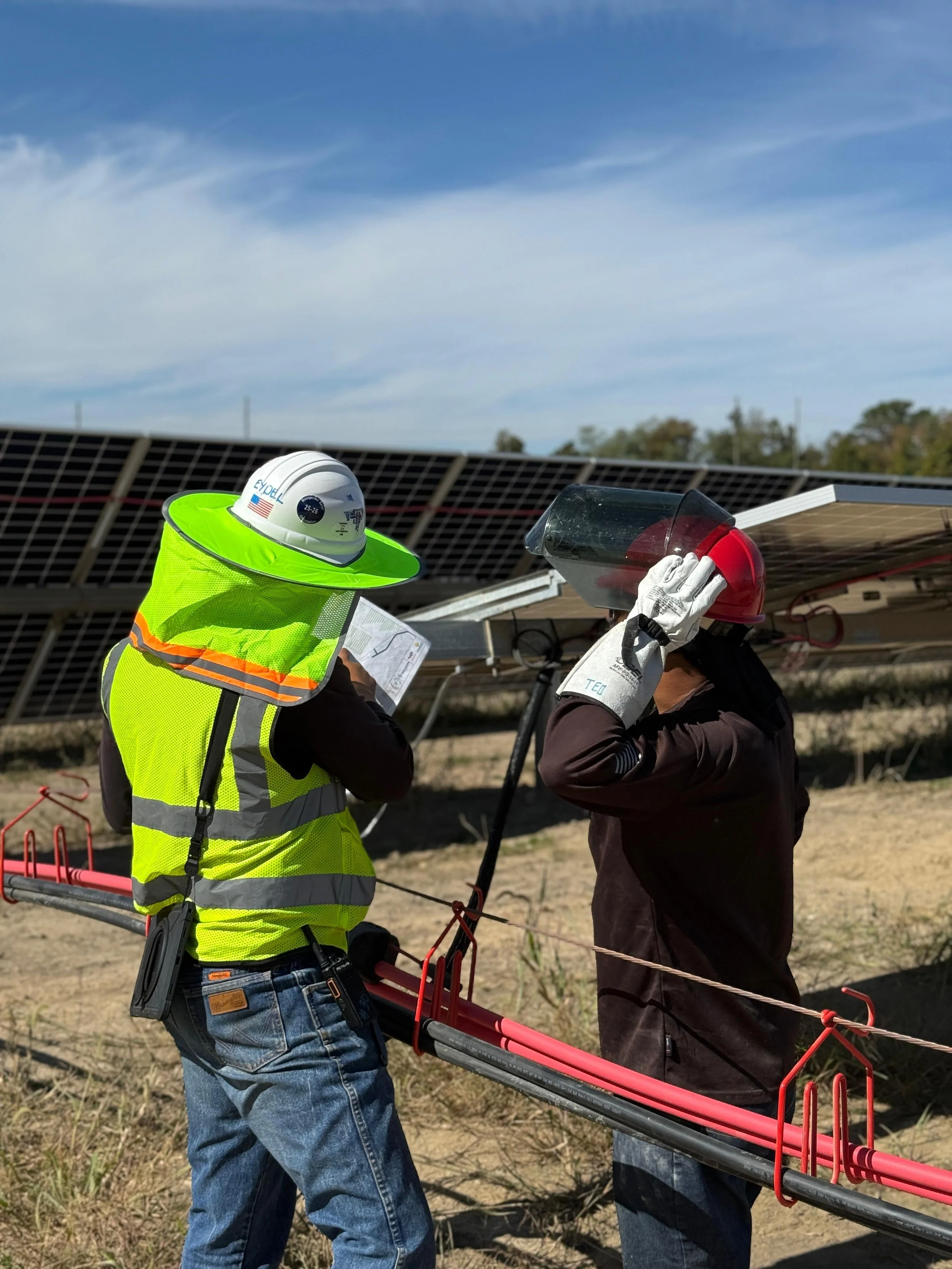 Two workers in safety gear inspecting under solar panels outdoors.