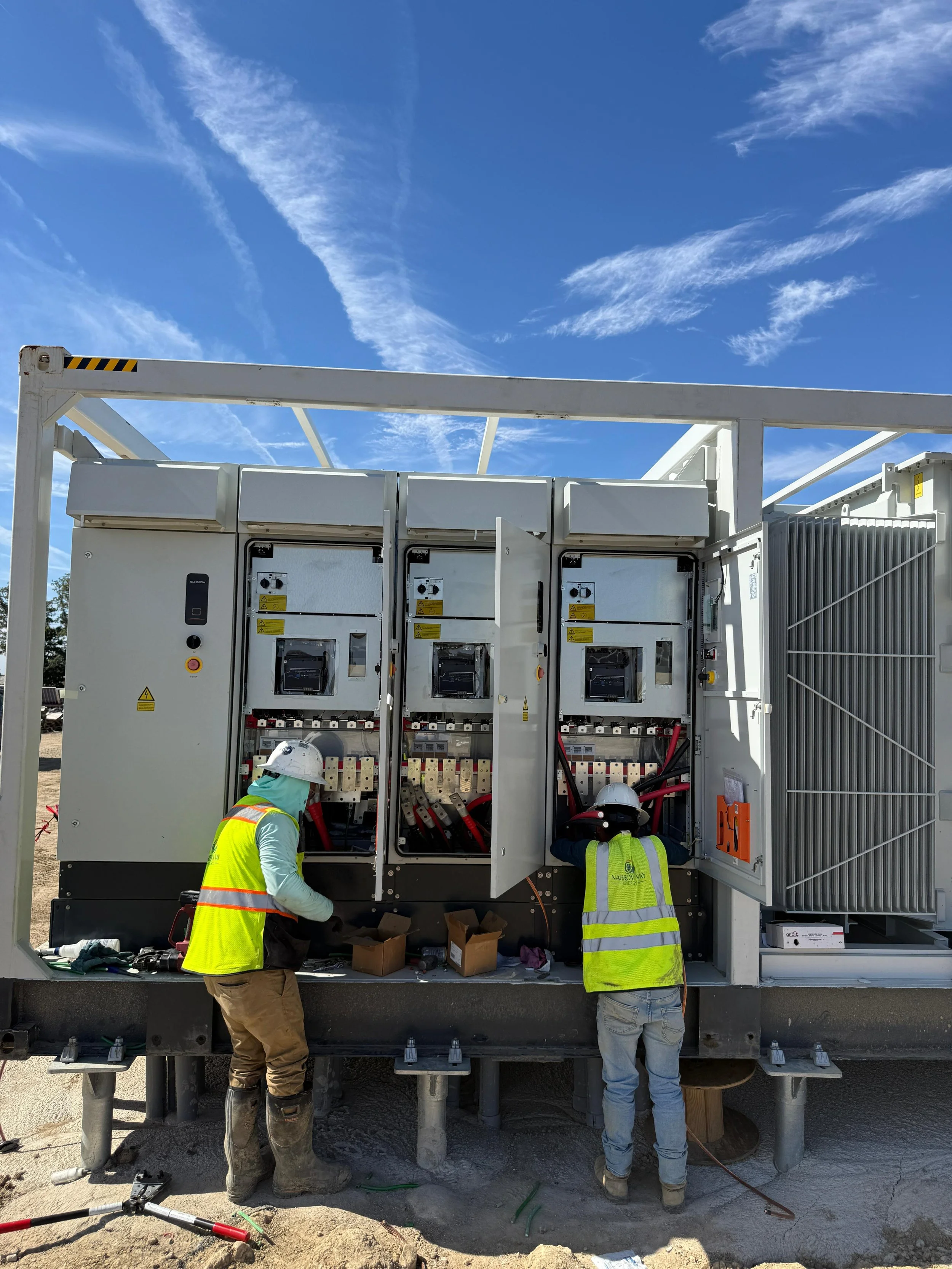 Two construction workers working on an electrical panel at a construction site, wearing safety vests and helmets, with a blue sky and clouds in the background.