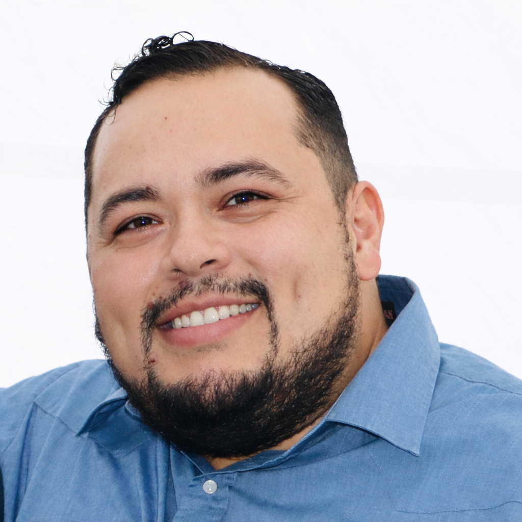 A smiling man with dark hair and a beard wearing a blue collared shirt.