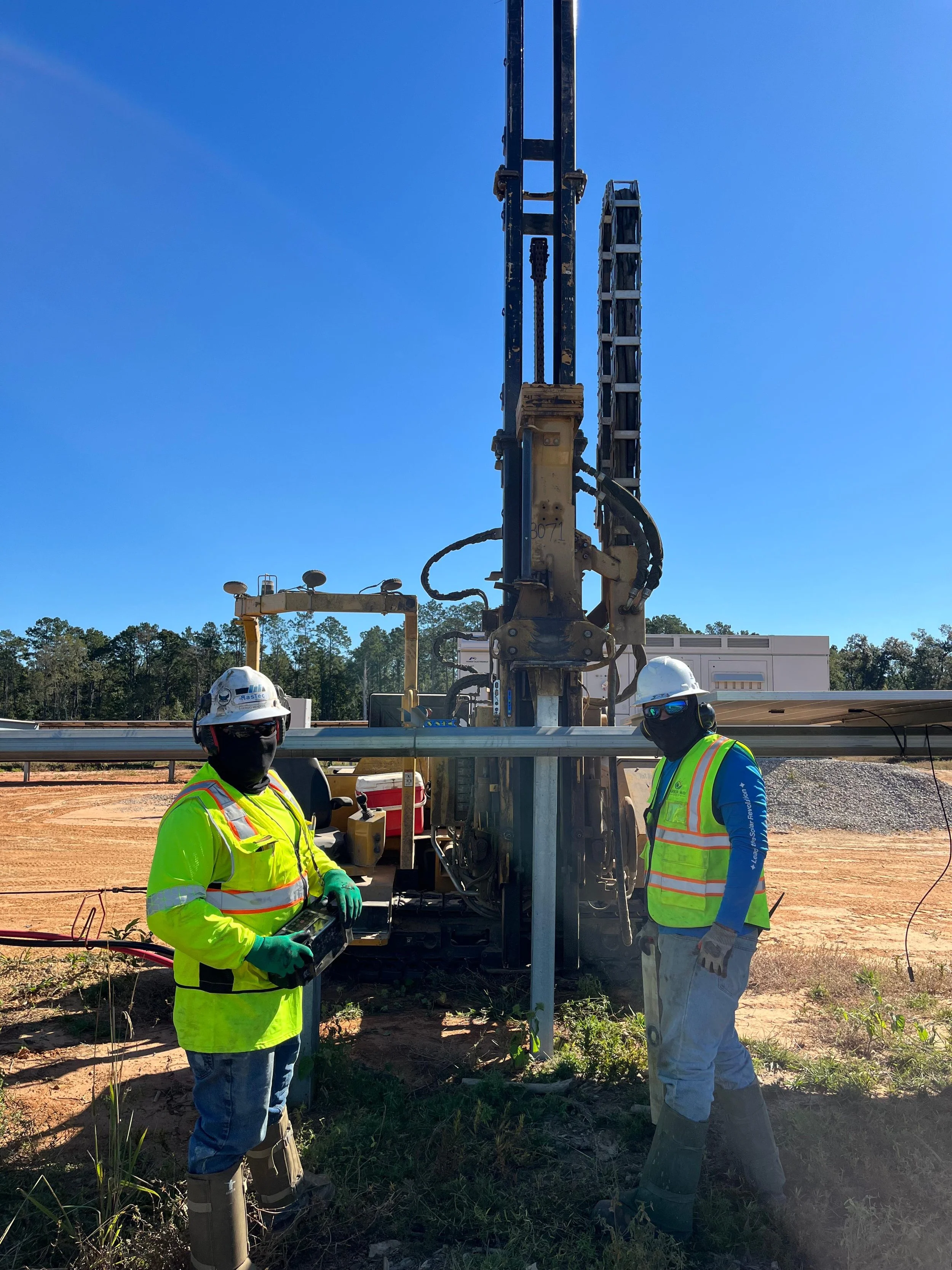 Two construction workers wearing safety gear and high-visibility vests operating a large pile driver at a construction site under a clear blue sky.