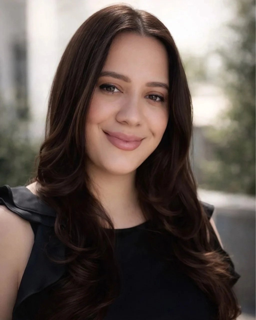 Close-up portrait of a smiling young woman with long, wavy brown hair, wearing a black sleeveless top, outdoors with blurred trees in background.
