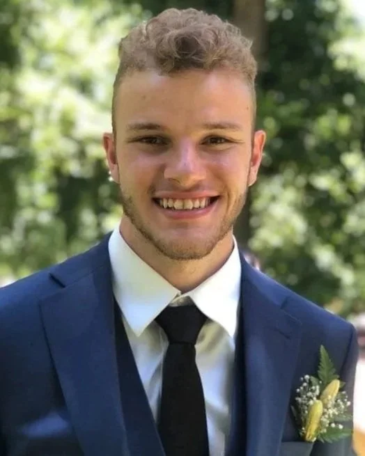 Young man smiling in a blue suit, white shirt, black tie, with a boutonniere, outdoors with trees in background.