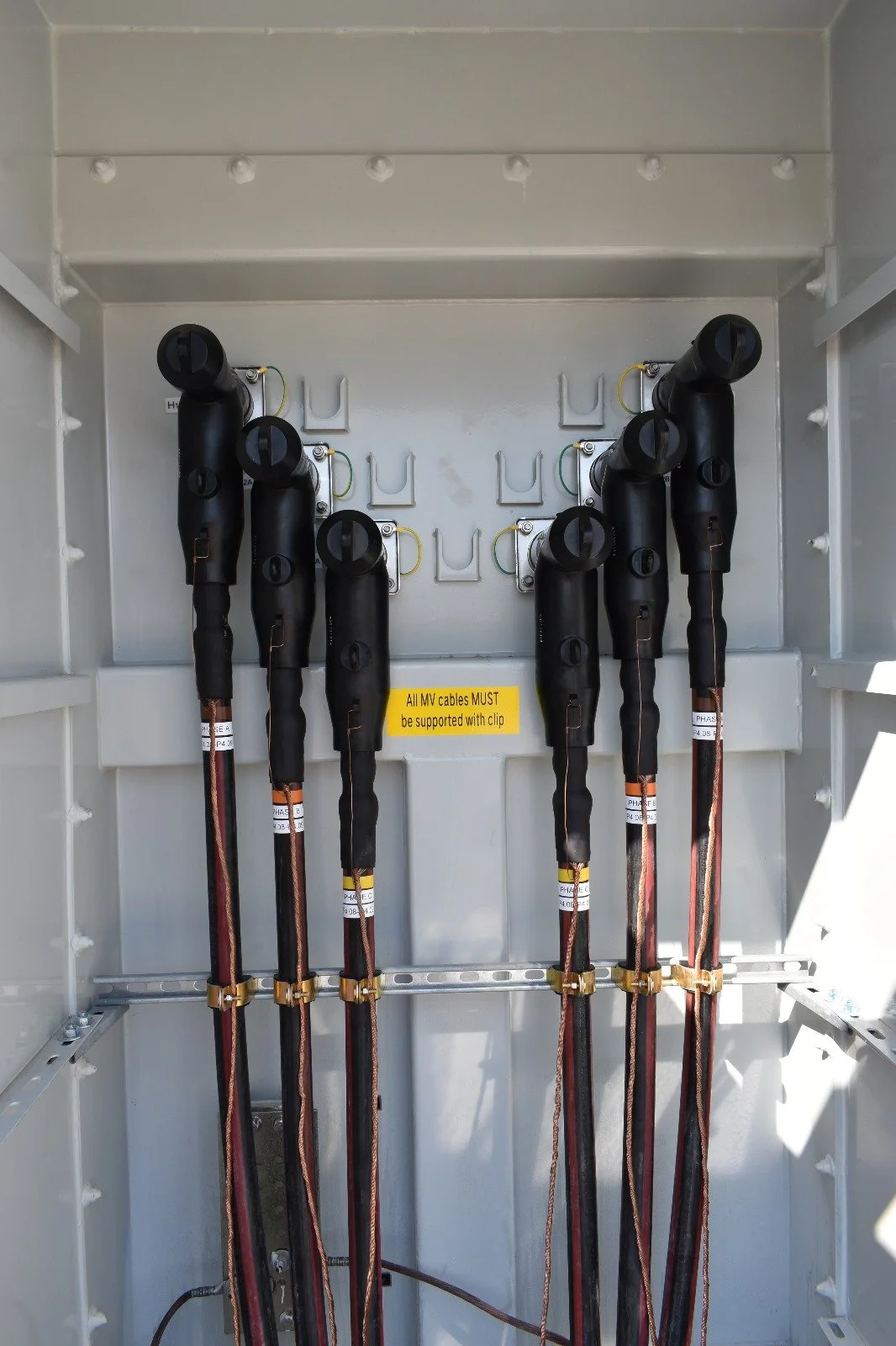 Electrical cabinet containing high voltage cables with a yellow warning sign that says, 'All MV cables MUST be supported with clip'.